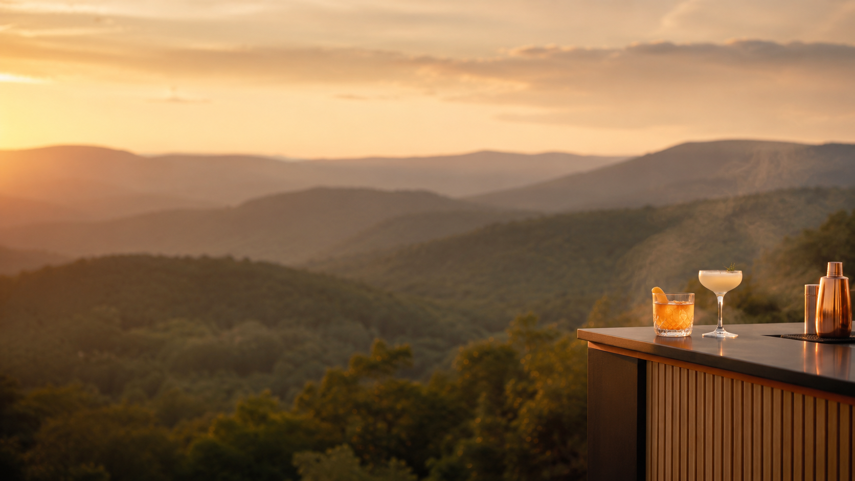 A bar counter with drinks and a copper cocktail shaker against a scenic mountain view at sunset.