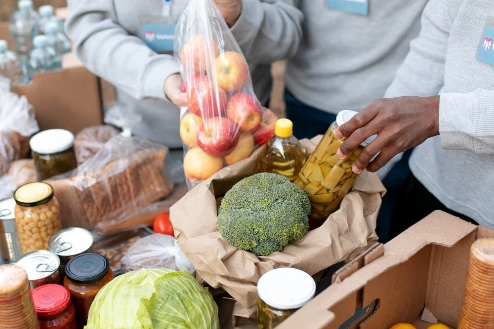 People packing groceries, including apples, broccoli, jars, and canned goods, into a paper bag.