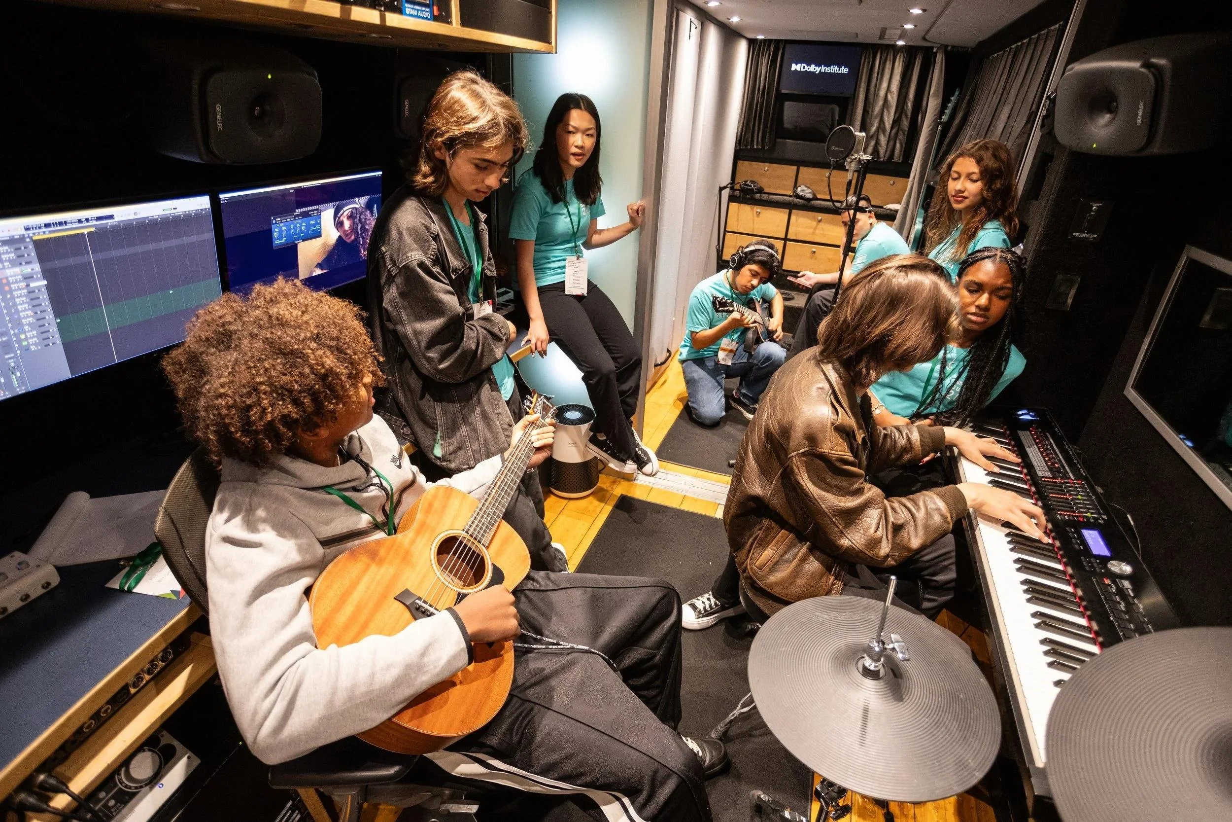 Group of young people in a recording studio, some playing guitars and a keyboard, others observing, with sound equipment and monitors around them.