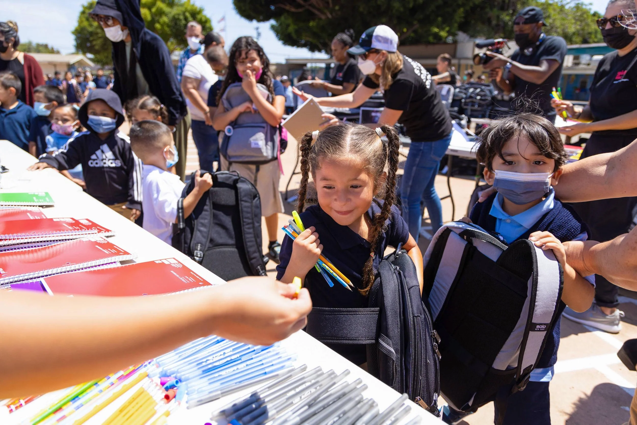 Children and adults wearing masks gather around a table outdoors with school supplies and notebooks, participating in an educational event.