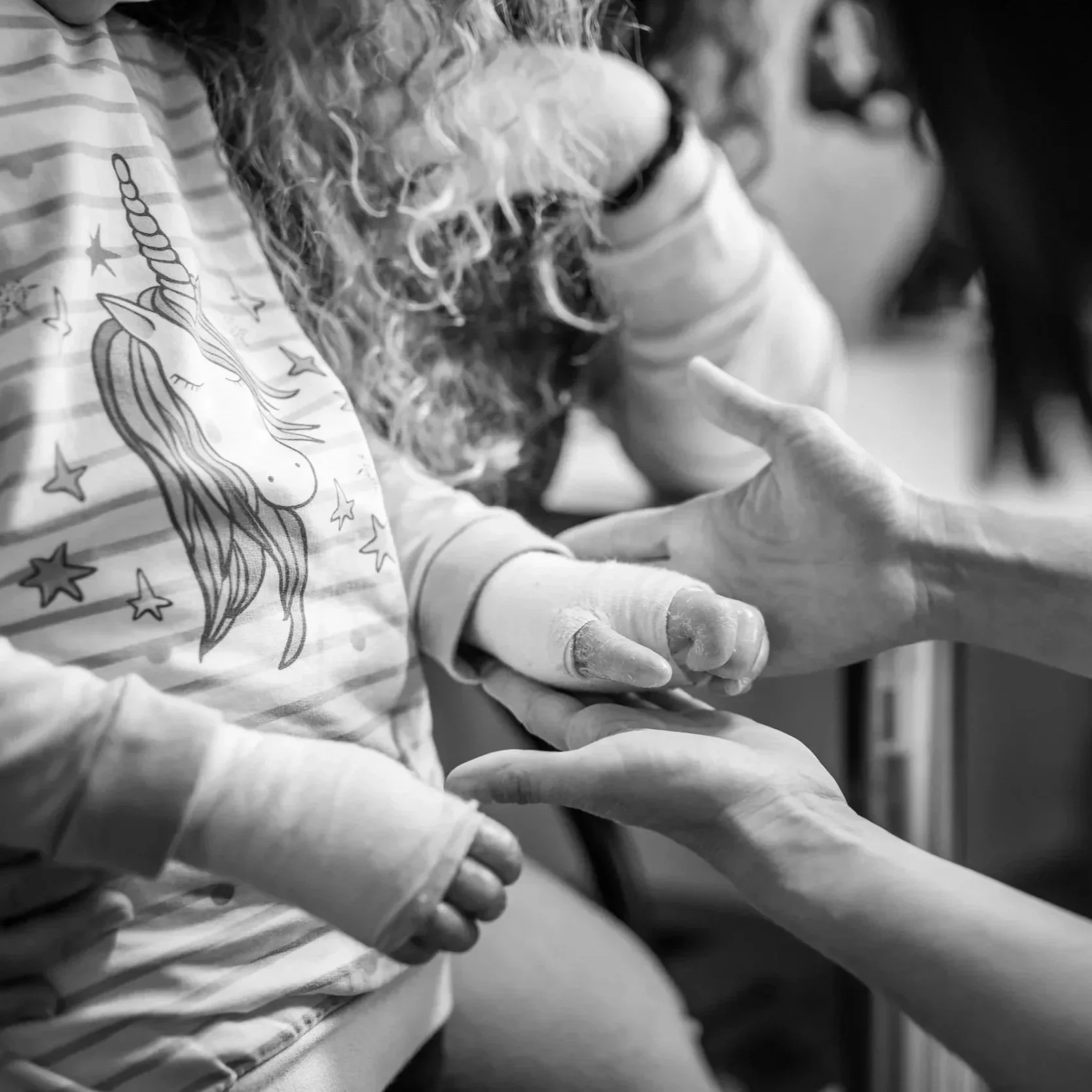 Child with curly hair and bandaged hands holding hands with an adult, wearing a unicorn shirt with stars.