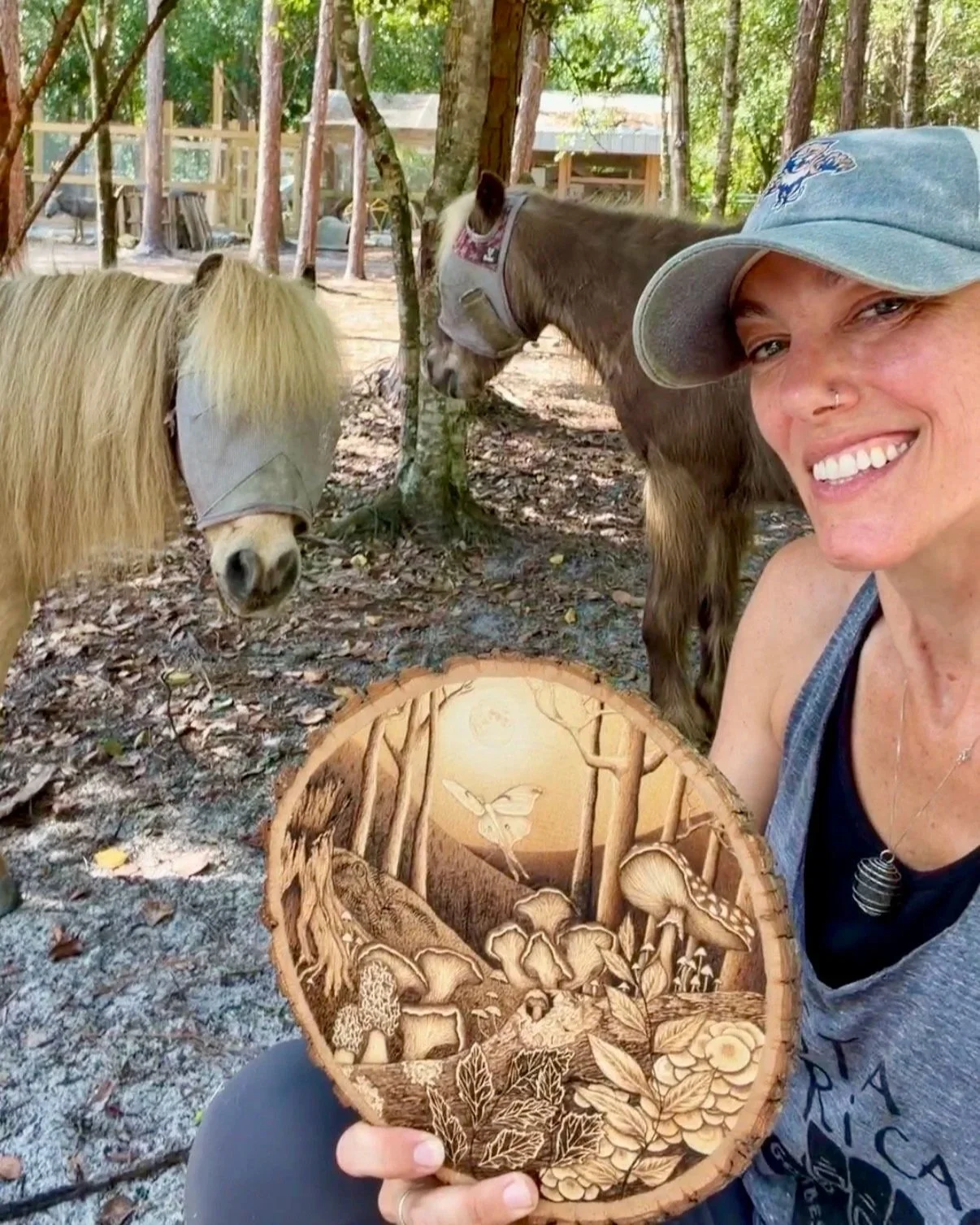A woman smiling in a forested area, taking a selfie with two horses in the background. She is wearing a blue cap and holding a carved wooden artwork depicting a forest scene with mushrooms, leaves, and a bird.