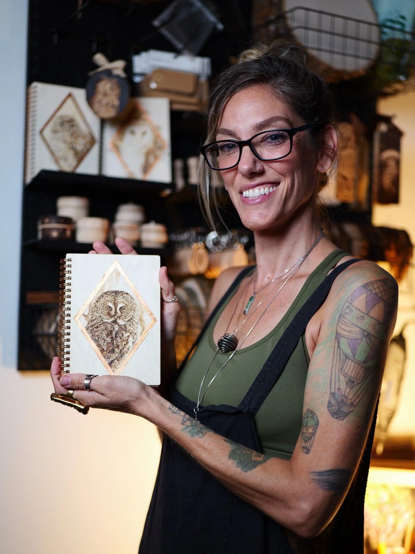A smiling woman with glasses, tattoos on her arms, and a nose ring, holding a small spiral-bound sketchbook featuring a detailed drawing of an owl, in a shop with art and craft supplies in the background.