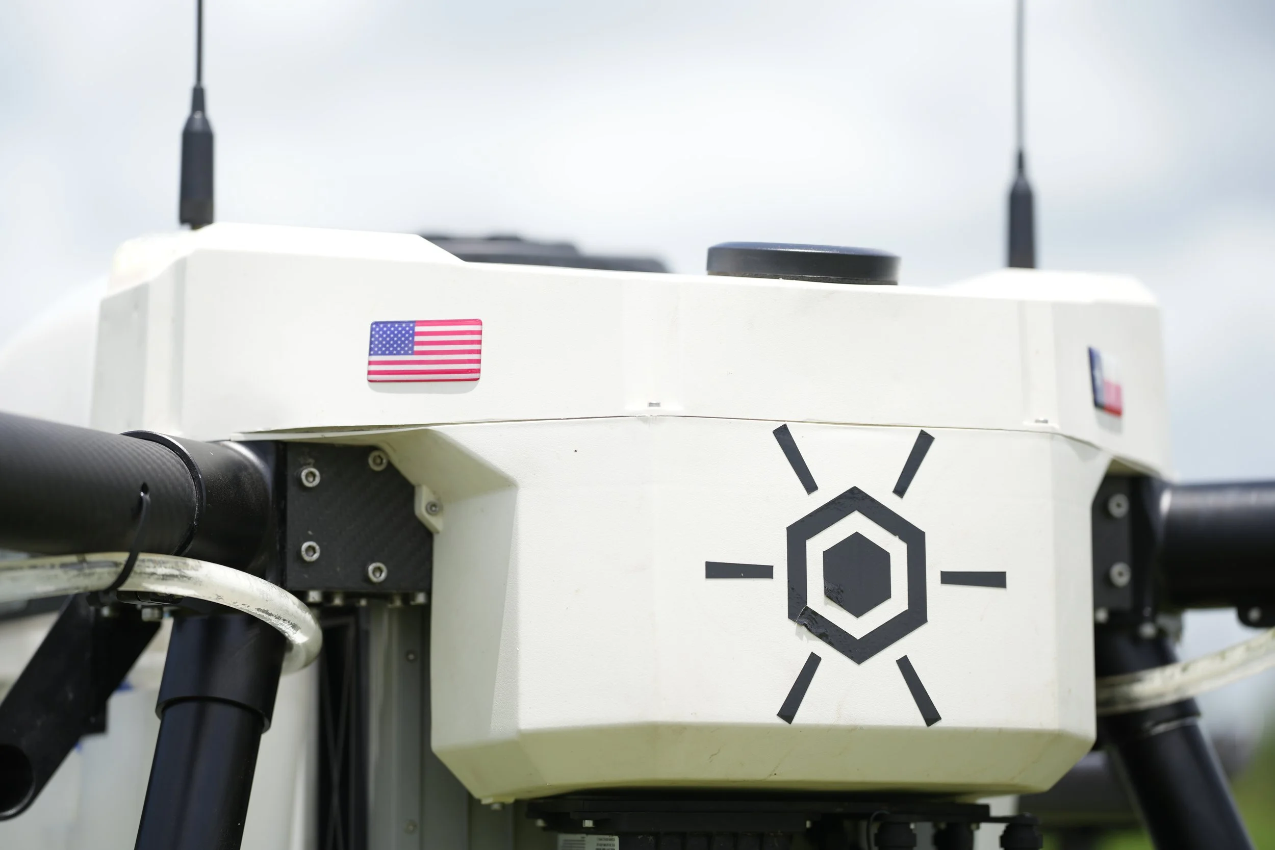 Close-up of a white robotic device with a small American flag sticker and a logo, featuring black and white components and antennas, on a light background.