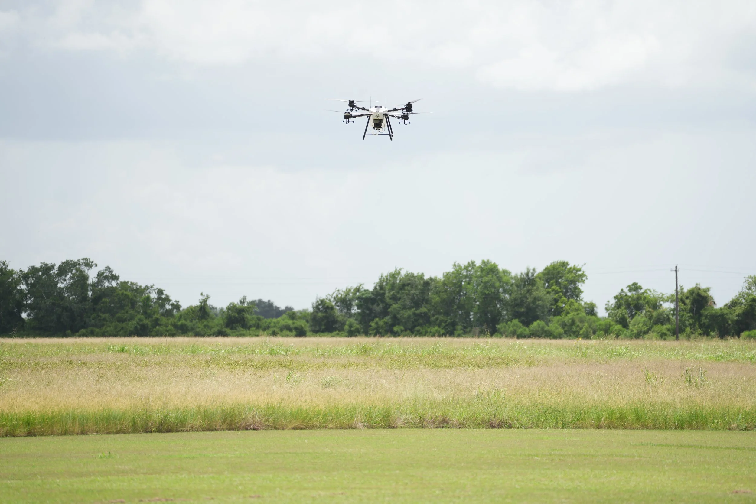 A drone flying in the sky over a grassy field with trees in the background and cloudy weather.