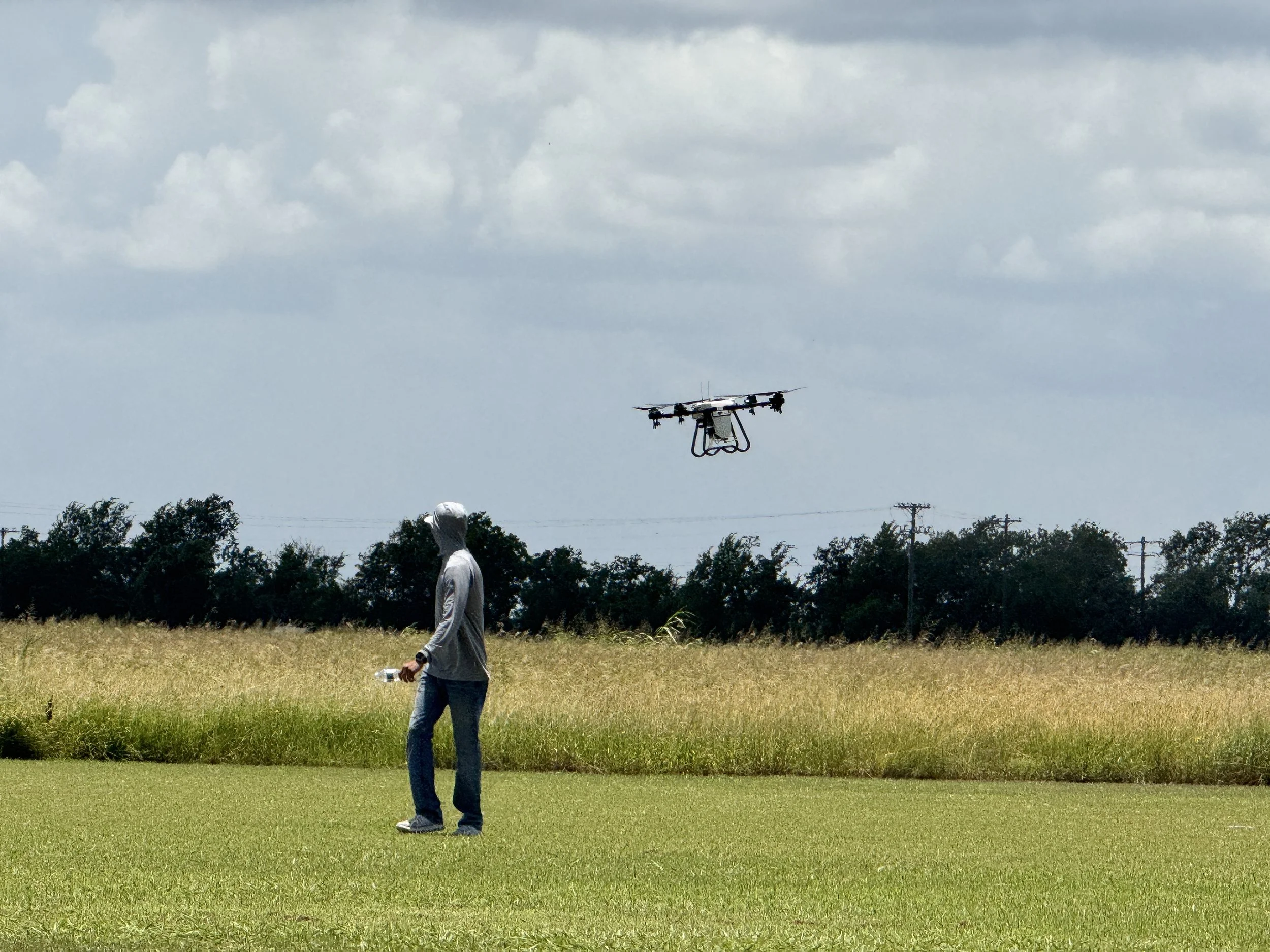 A person in a gray hoodie and jeans walking in a grassy field holding a water bottle, with a drone flying in the sky overhead and trees in the background.
