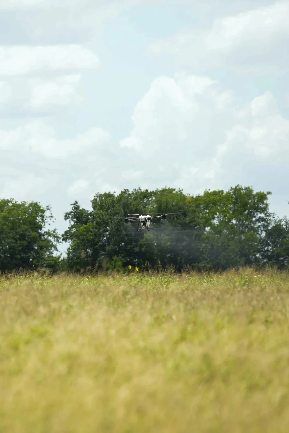 A drone flying over a grassy field with trees in the background and a cloudy sky above.