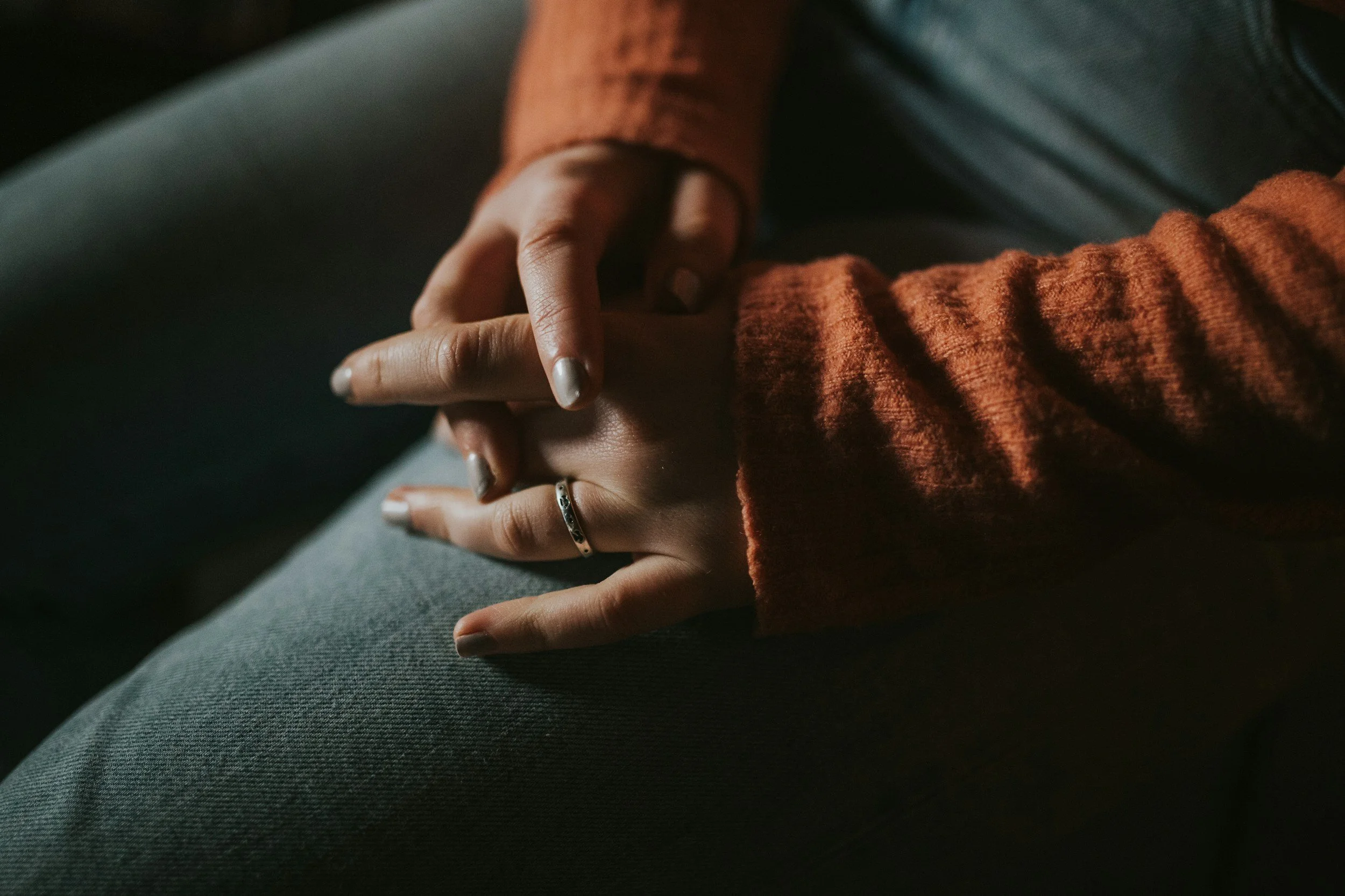 A person wearing a brown sweater and gray pants is sitting with their hand resting on their knee, showing silver rings on their fingers, with radiant natural lighting highlighting their hand and textured fabric.