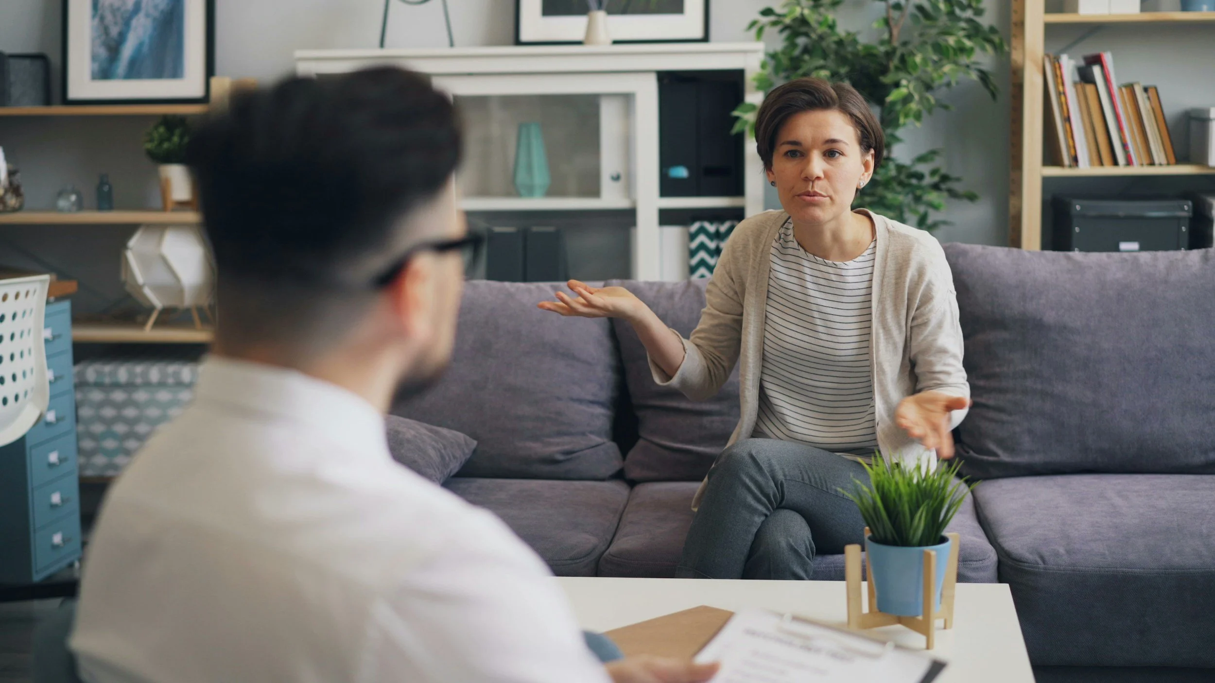 Two people having a discussion in a cozy living room, with shelves, framed pictures, plants, and a gray couch.