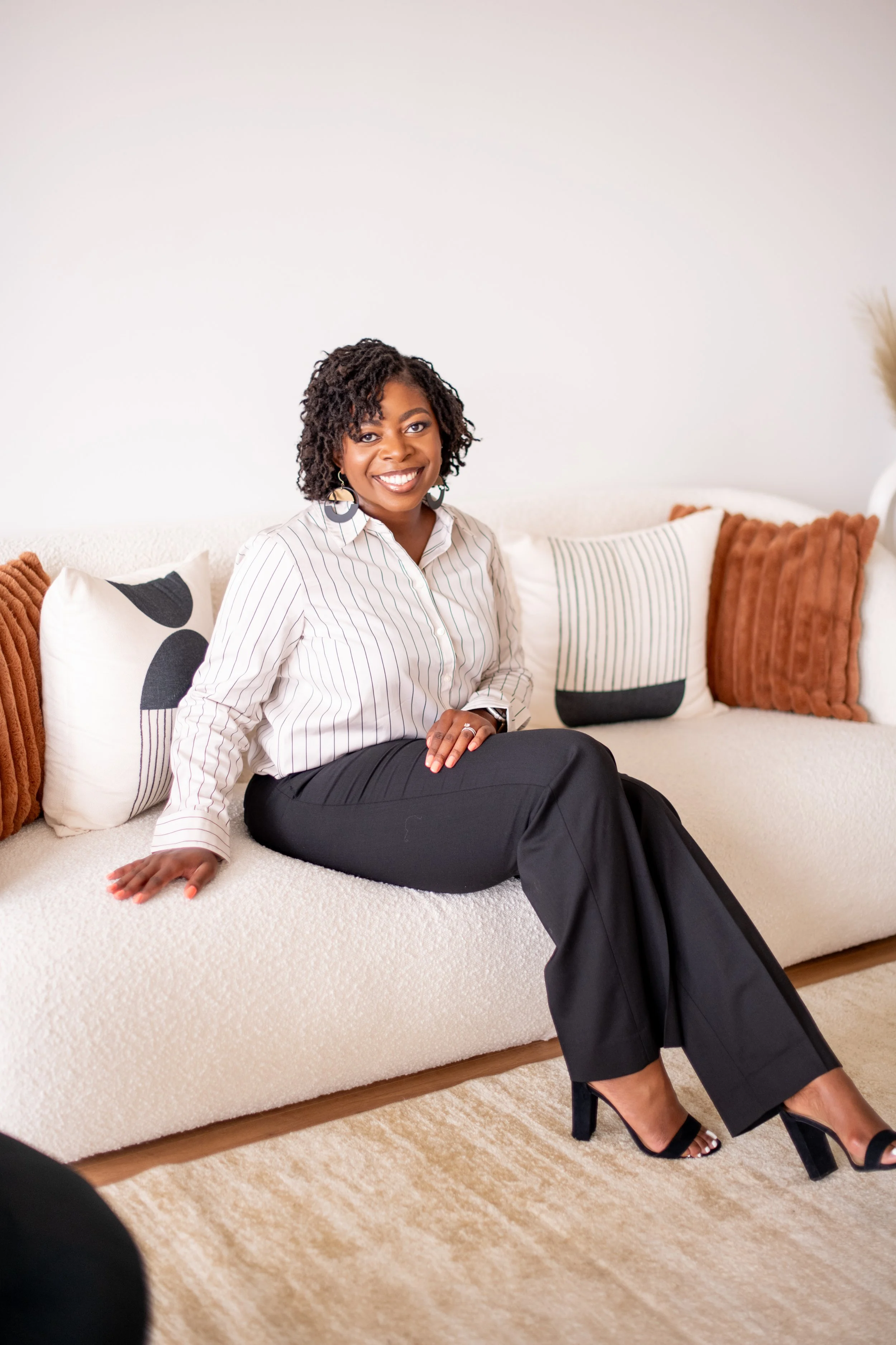 A woman sitting on a cream-colored sofa with decorative pillows, smiling at the camera in a room with a beige carpet and white wall.