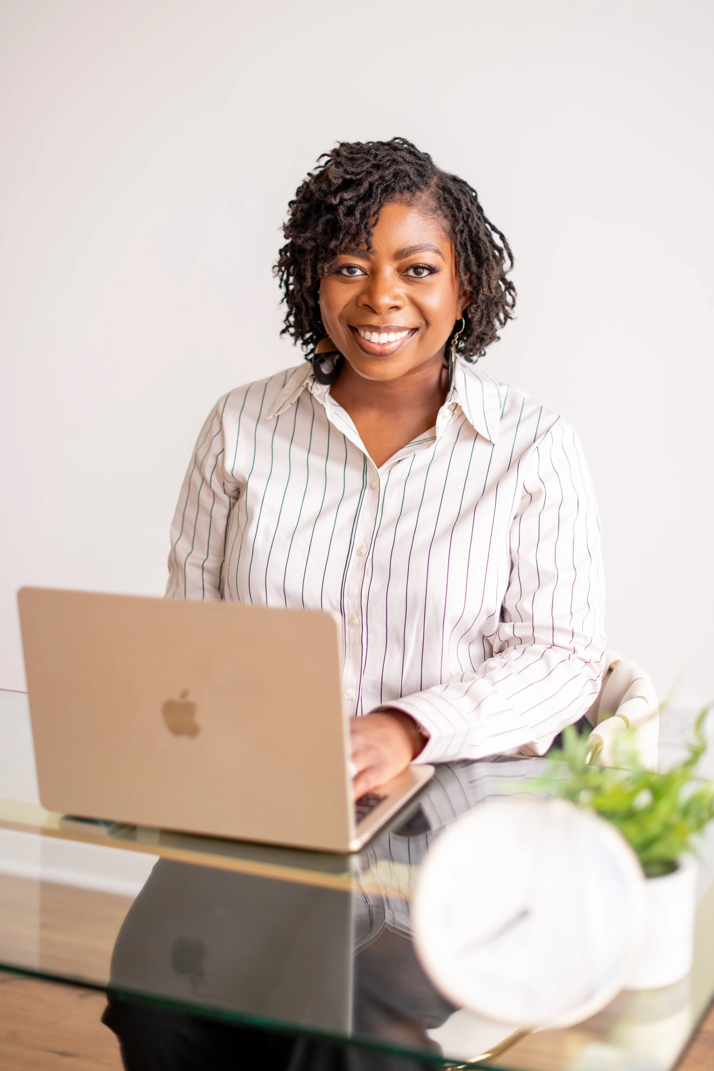 A woman with mircolocs smiling while working on a silver MacBook at a glass table, with a plant and a white globe in the foreground.
