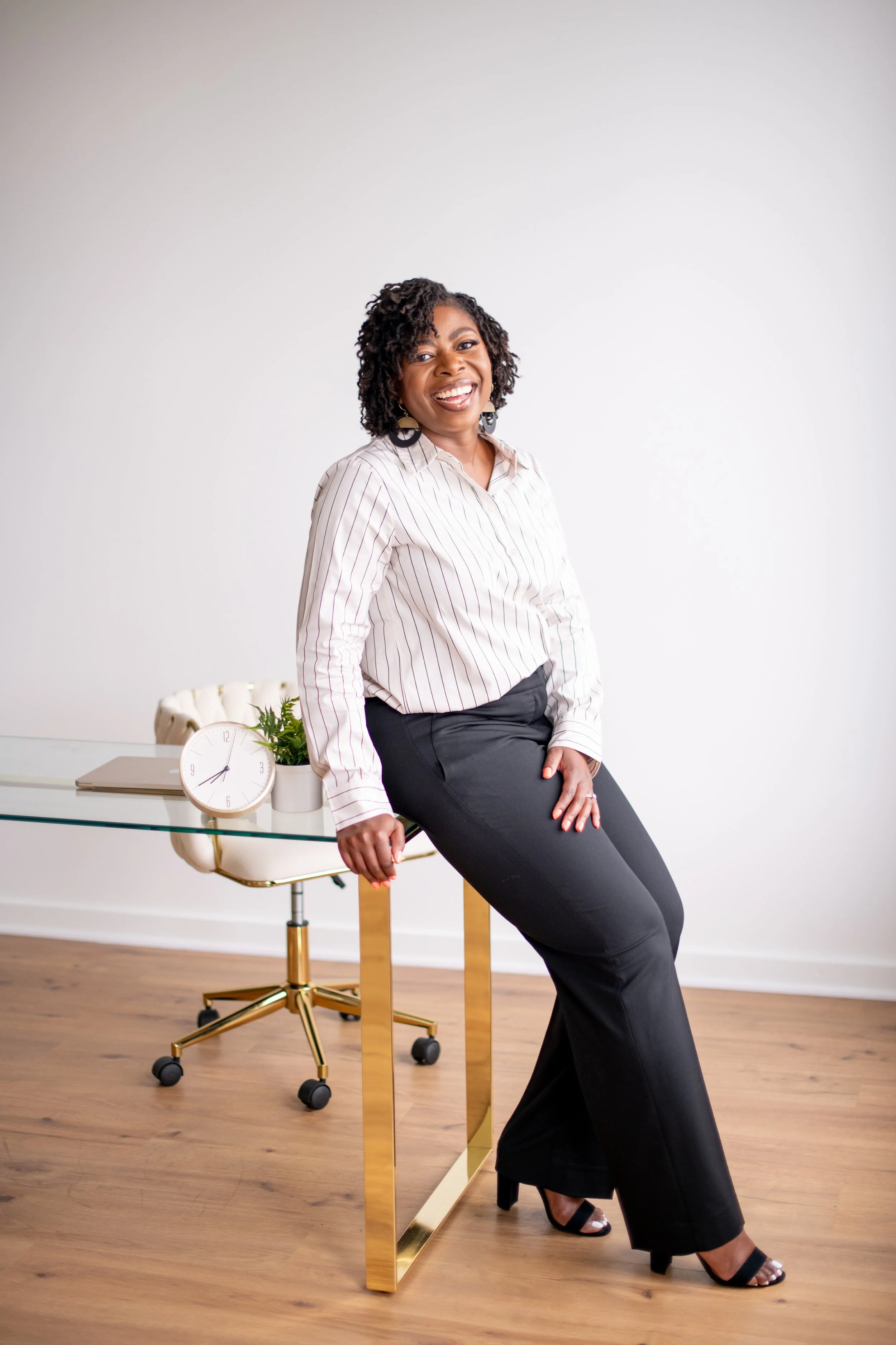 A woman with mircolocs wearing a striped shirt and black pants, sitting on a table in a modern office. Behind her there is a glass table with a white clock, a notebook, and a plant, and a white office chair with gold accents.