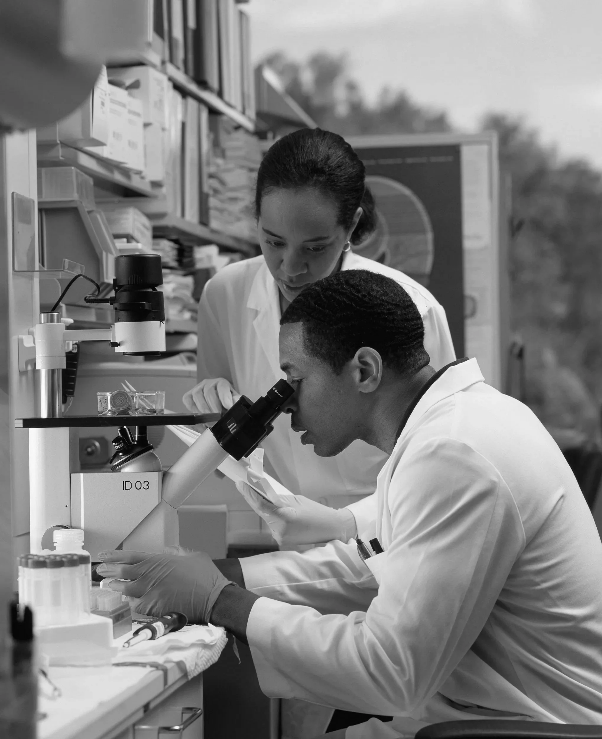 Two scientists in white lab coats working together in a laboratory, examining samples through a microscope, with shelves of laboratory supplies in the background.