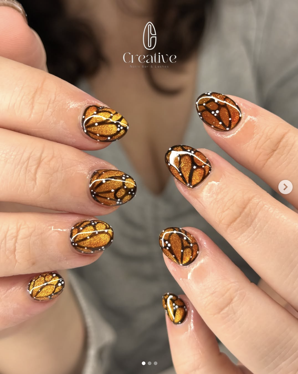 Close-up of a person showing off the nails decorated with orange, black, white, and gold butterfly wing-inspired nail art design at a nail salon.