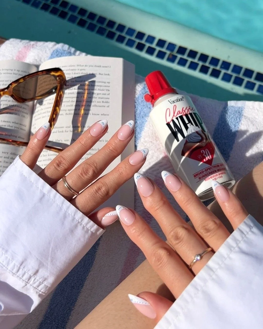 Person with manicured nails near a swimming pool, holding a bottle of sunscreen, with sunglasses, an open book, and a folded towel nearby.