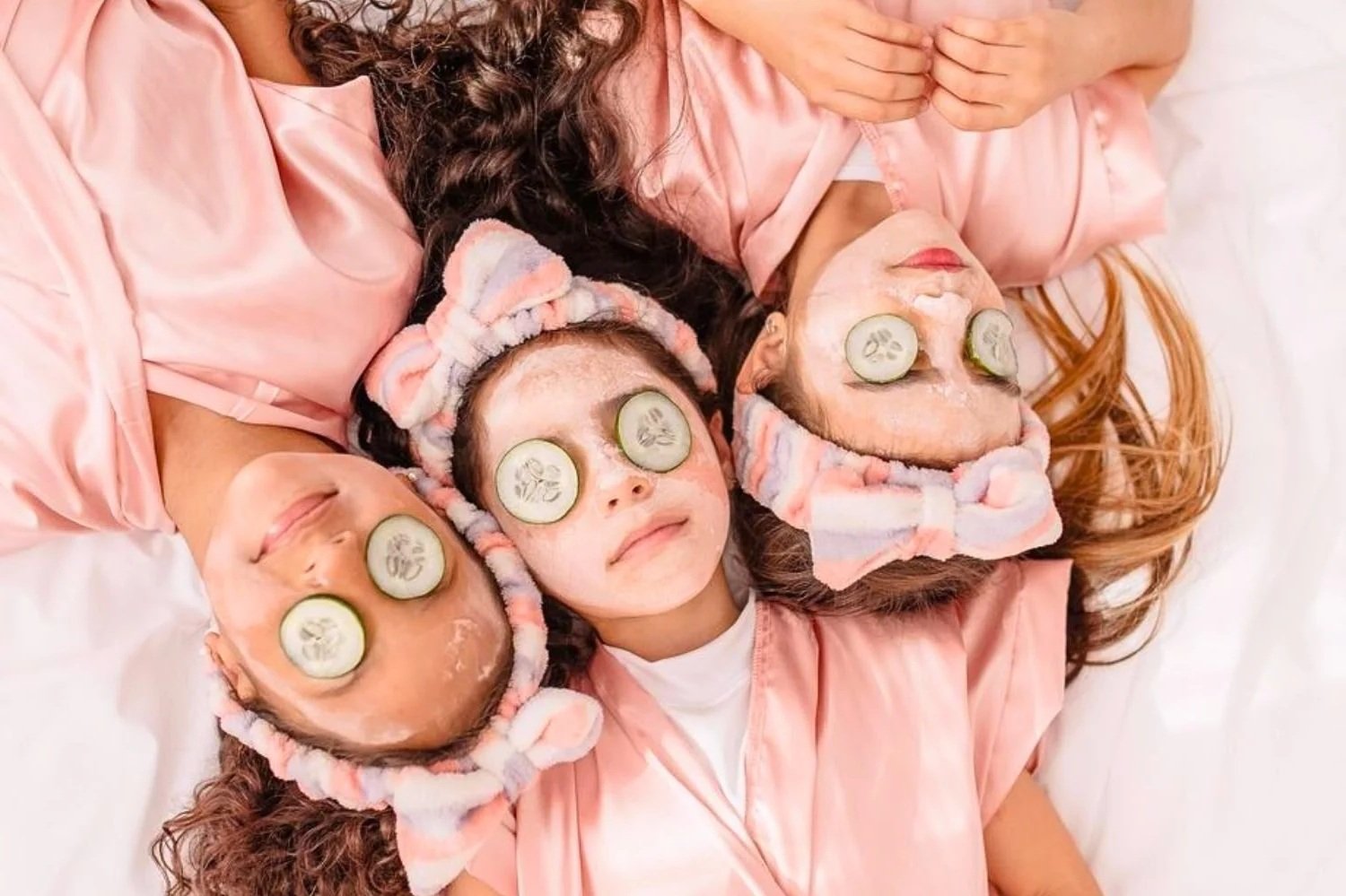 Three girls laying on a bed with cucumber slices over their eyes. They are wearing pink robes and have pink and white striped headbands.