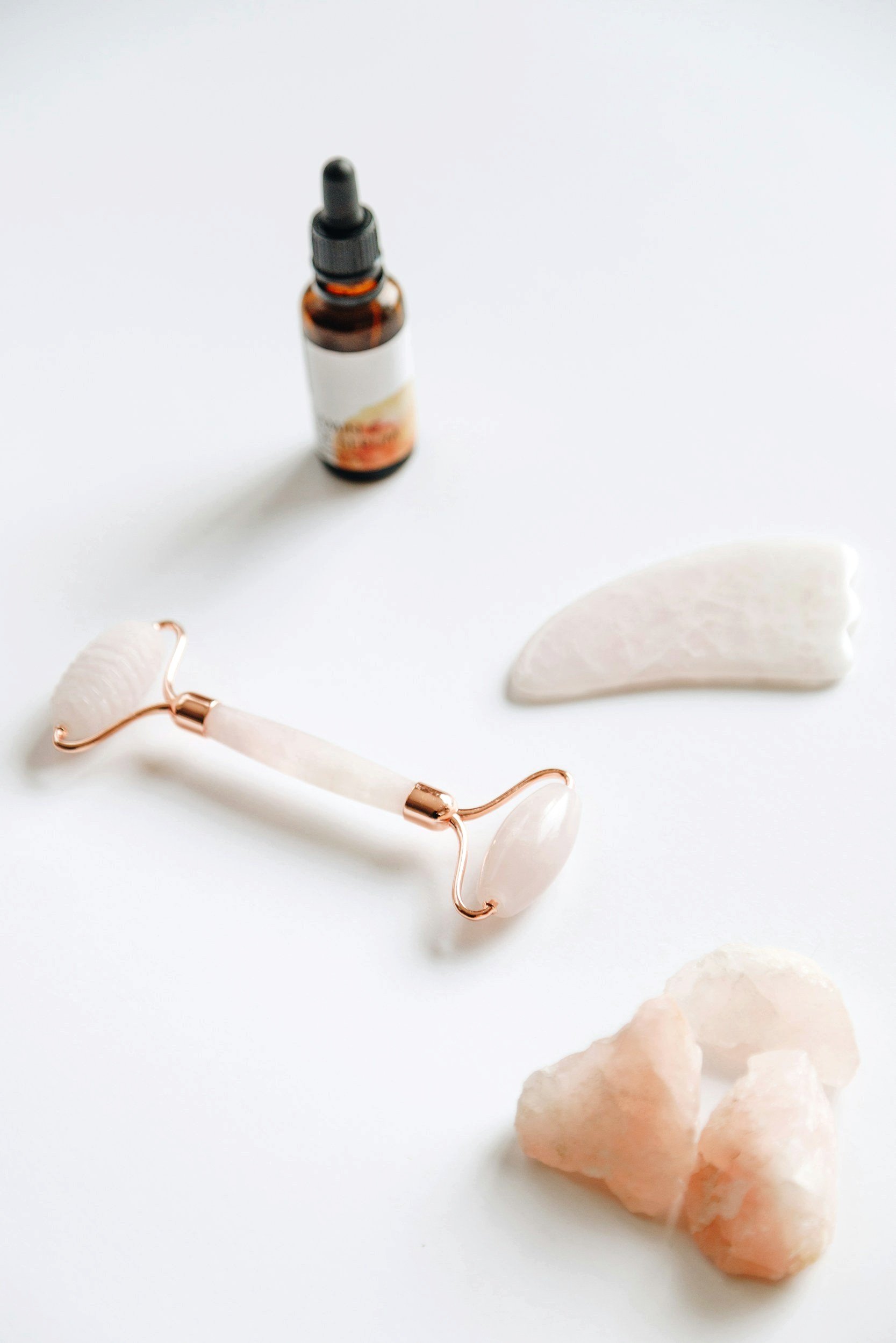 Small amber dropper bottle with a black cap and dropper, a white facial roller with rose gold accents, a light pink star-shaped crystal, and scattered light peach-colored stones on a white background.