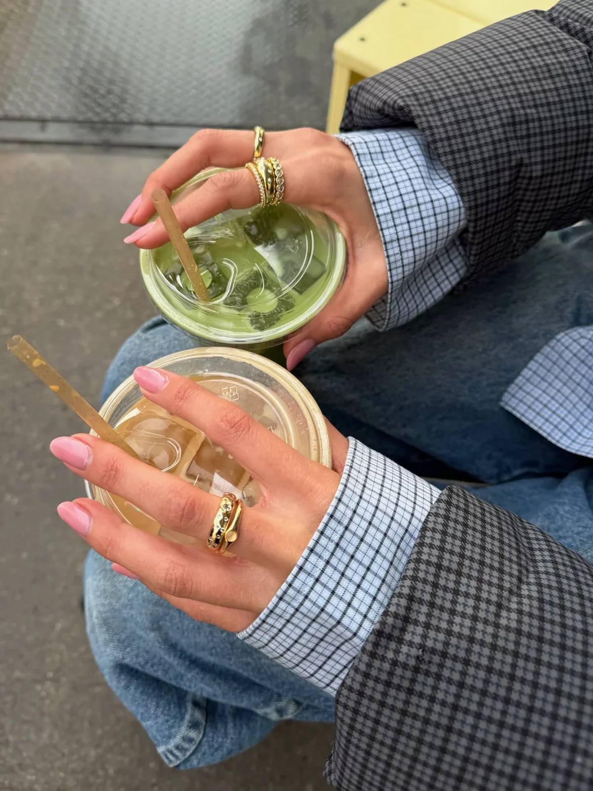 Two hands holding plastic cups with iced beverages, one green and one brown, with straws. The person has pink nail polish and wears rings, a plaid blazer, and a checkered shirt. They are sitting on a person's lap, with a yellow table in the background.