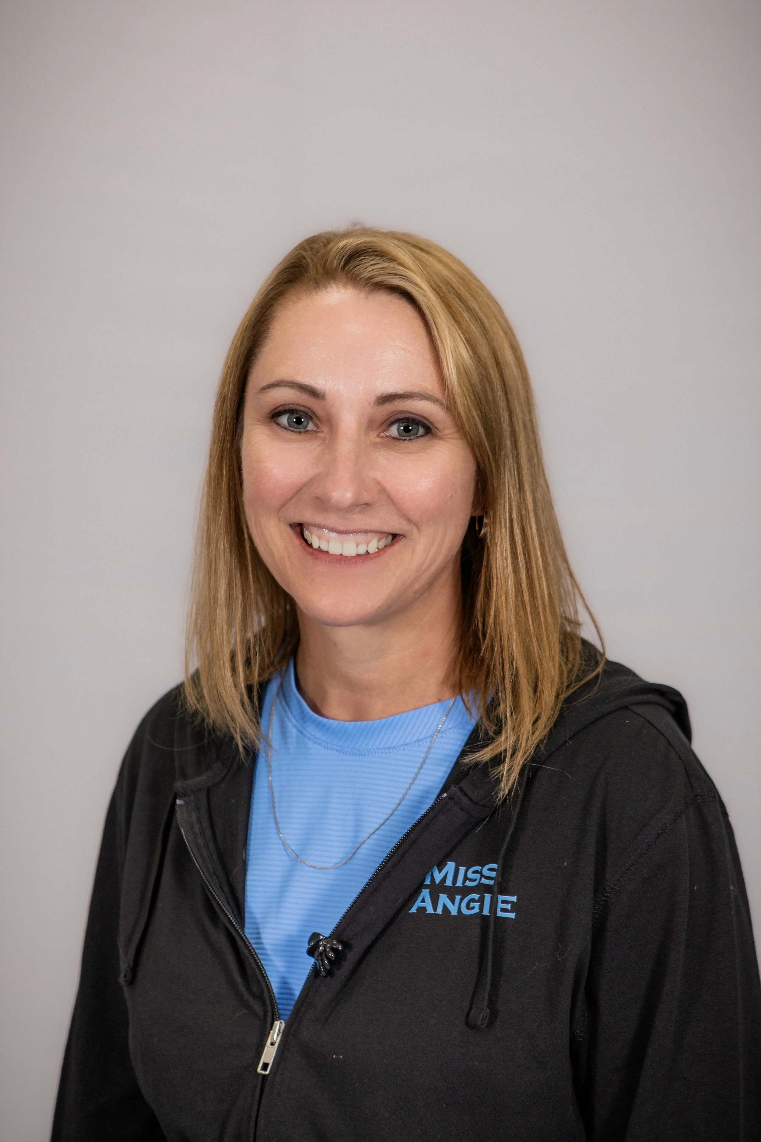 Portrait of a smiling woman with blonde hair, wearing a blue athletic shirt and a black jacket with a logo that reads 'Miss Angie,' standing against a plain gray background.
