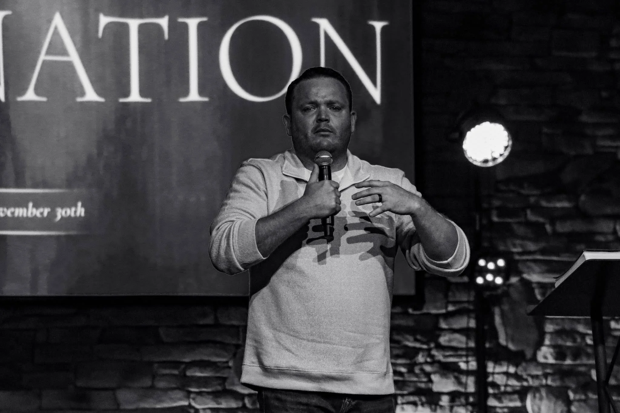 A man holding a microphone on stage in front of a background with the word 'ATTENTION' visible. He appears to be speaking or performing, with a serious facial expression.