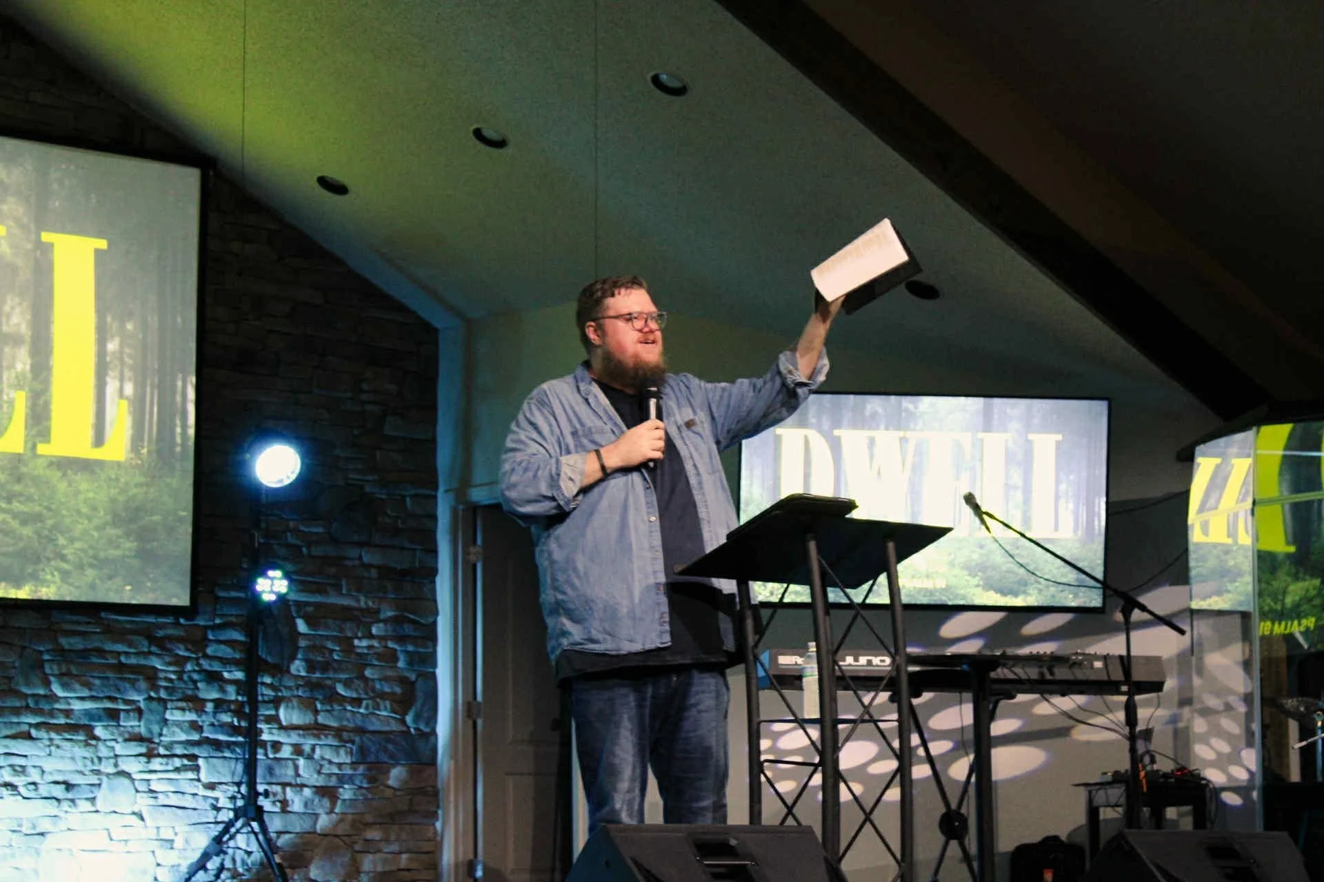 Man with glasses and a beard, holding a microphone in one hand and a booklet in the other, speaking at a presentation or event. There are two large screens behind him displaying the word 'DWELL' with a forest background, and stage lighting is visible.