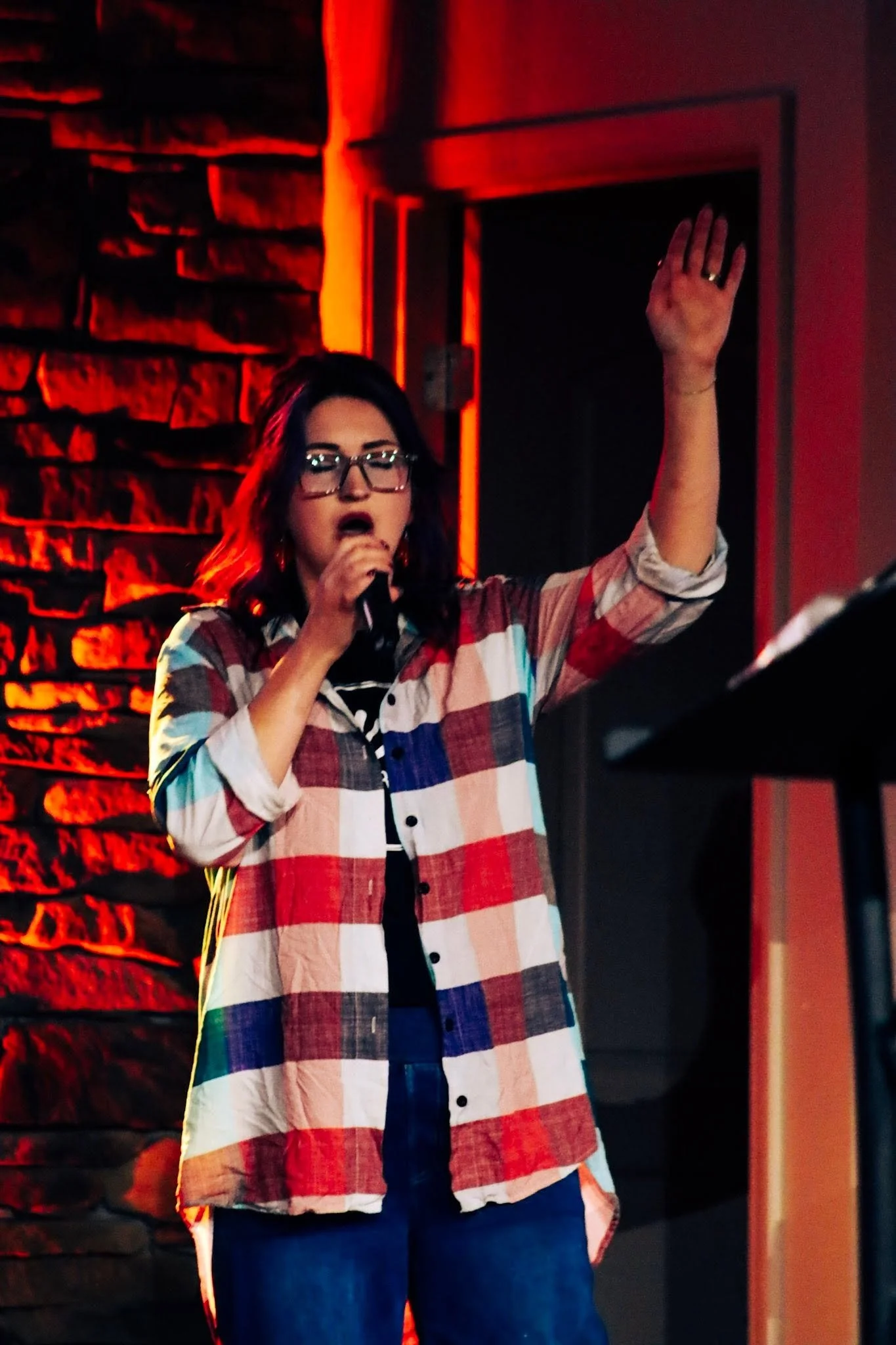 A woman in glasses holding a microphone, raising one hand, wearing a plaid shirt, and standing in front of a brick wall with red lighting.