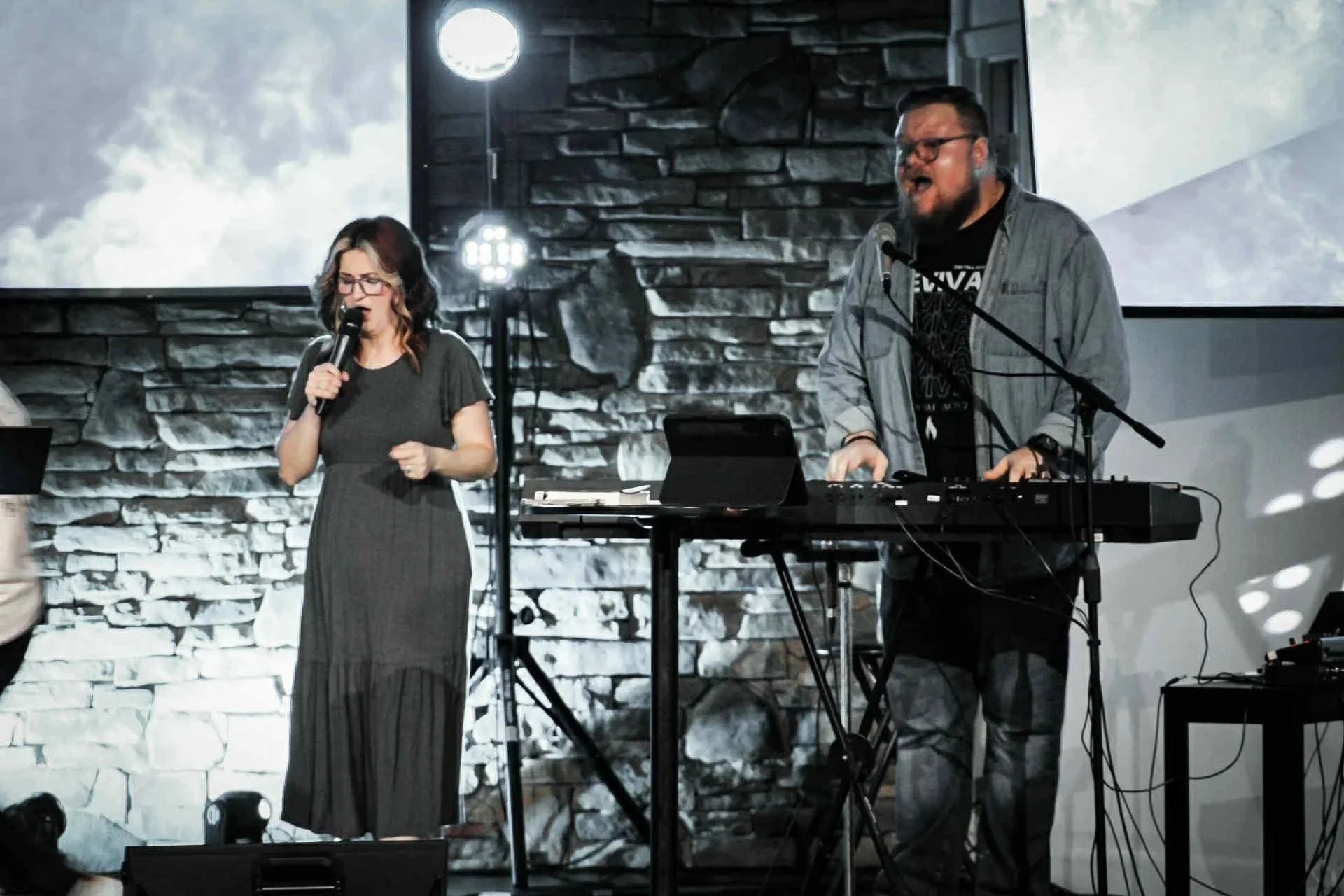A woman singing into a microphone and a man playing a keyboard on stage with a stone wall background and large screen overhead.