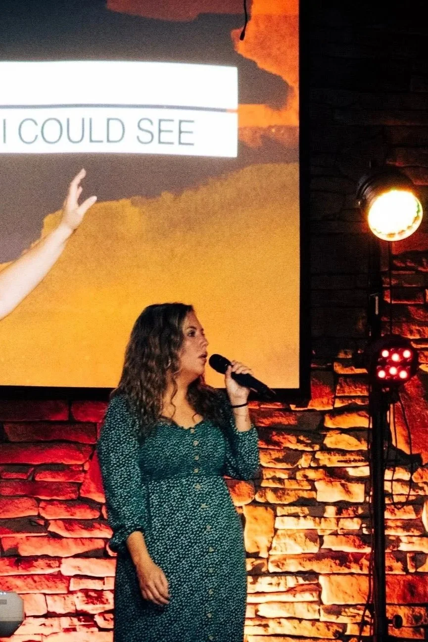 A woman with curly hair and a patterned dress holding a microphone, standing in front of a brick wall with red lighting.