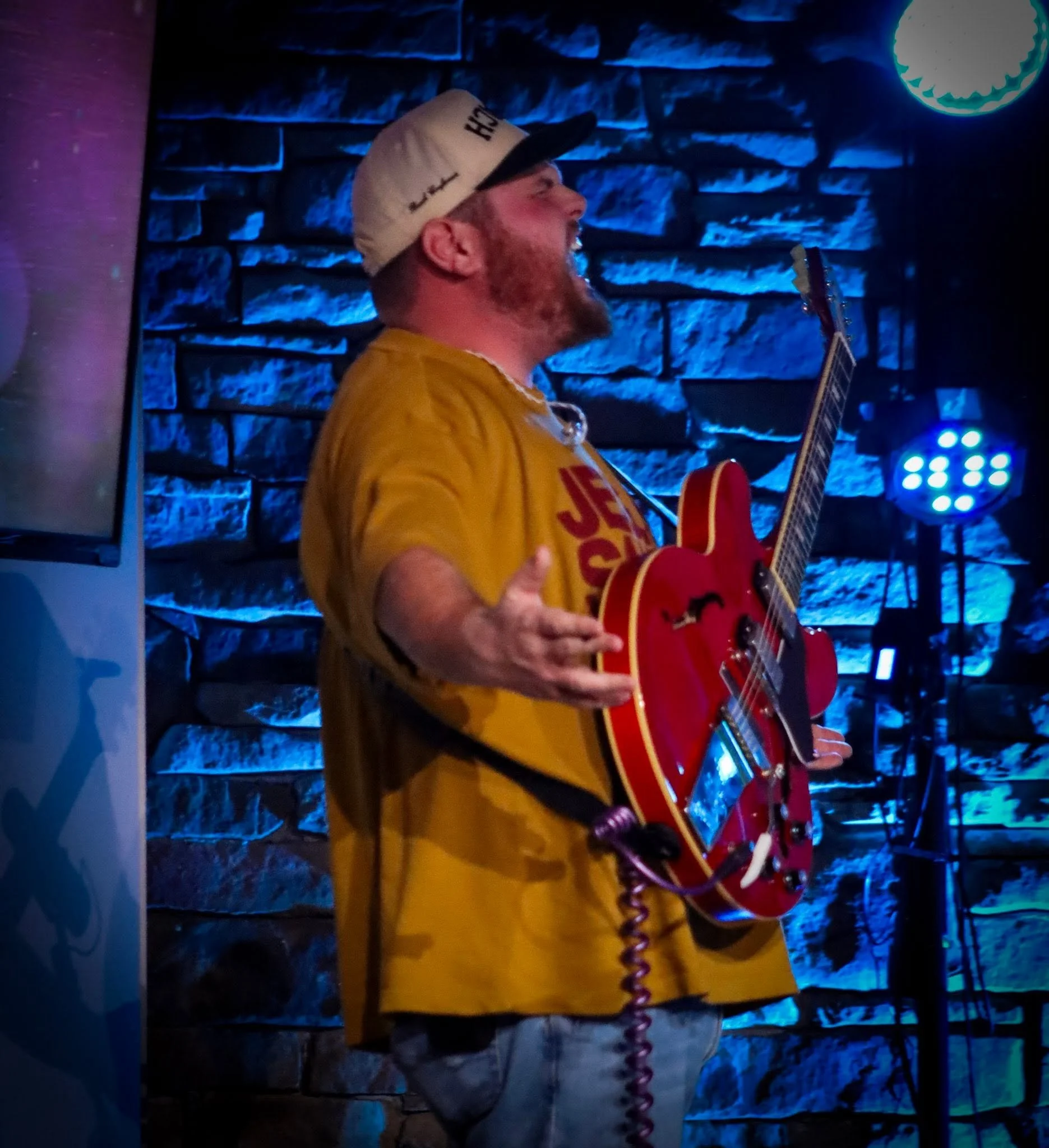 A man with a red beard and a beige cap playing an acoustic guitar in a dimly lit room with a dark stone wall illuminated by blue lights in the background.