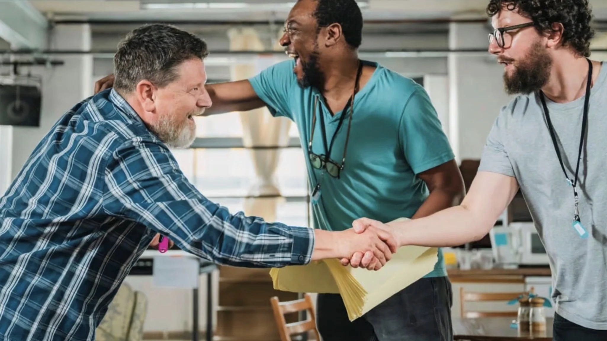 Three men smiling and shaking hands in an office or workshop setting, one with a beard wearing a plaid shirt, another wearing glasses and a gray t-shirt, and the third with a dark beard wearing a blue t-shirt.