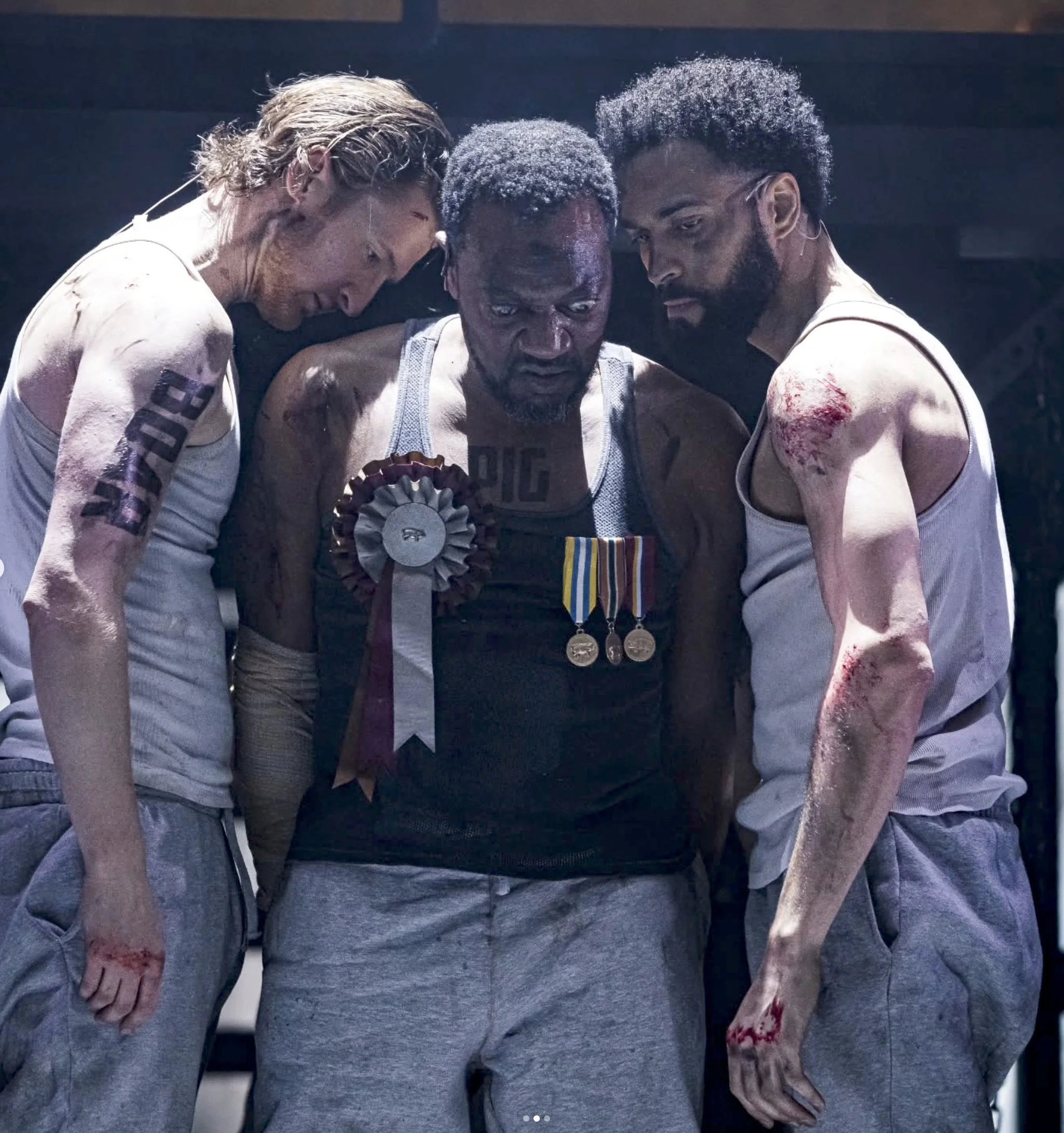 Three men, who all appear to be injured with blood and cuts on their bodies, are standing close together with somber and intense expressions on their faces. They all have different hairstyles and are wearing sweaty tank tops or shirts, with medals ha
