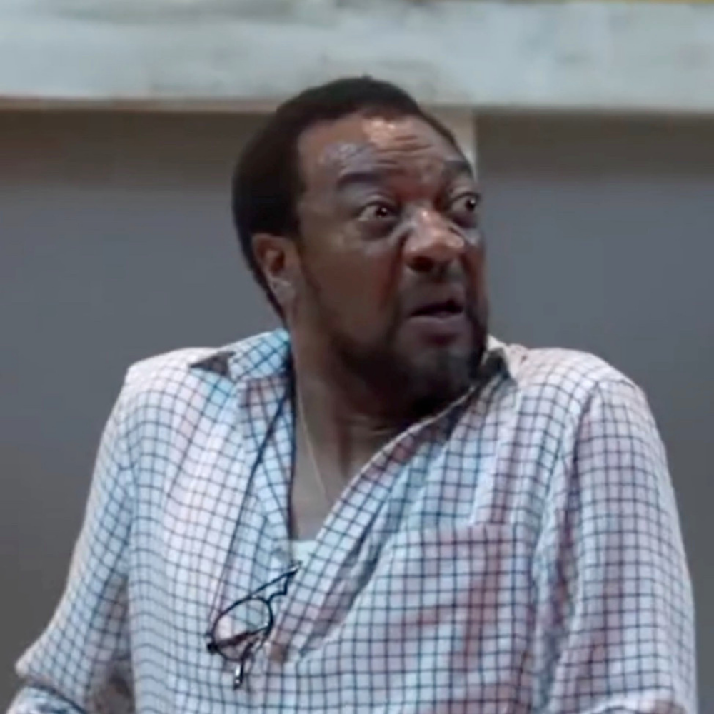 A man with a surprised or angry expression, wearing a checkered shirt, sitting indoors in front of a gray wall.
