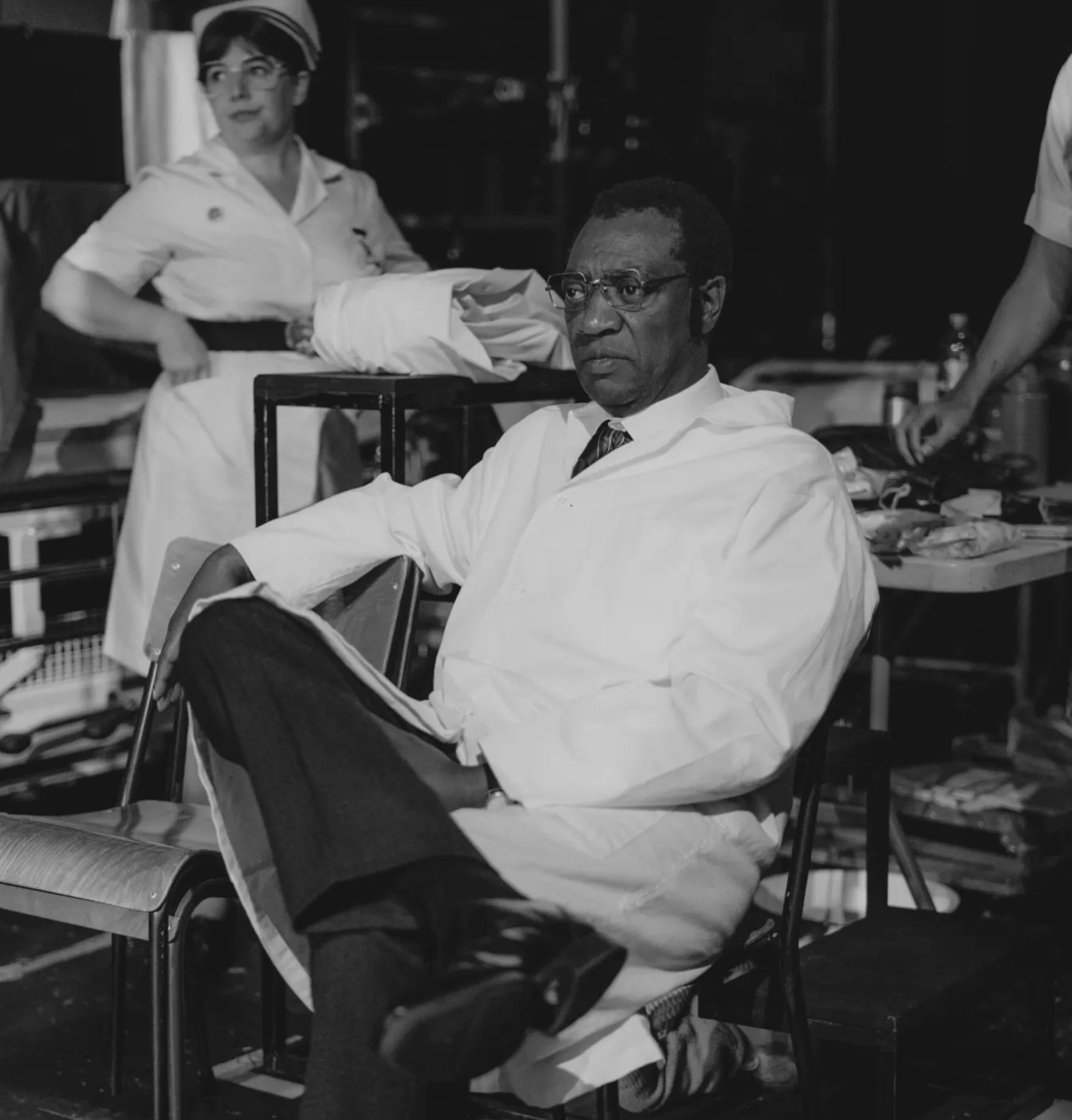 Black and white image of Fred Rogers and an African American man with glasses sitting in a room with chairs and a cart filled with various items. Fred Rogers is wearing a tie and a white shirt, while the man appears to be in a white shirt and dark ve