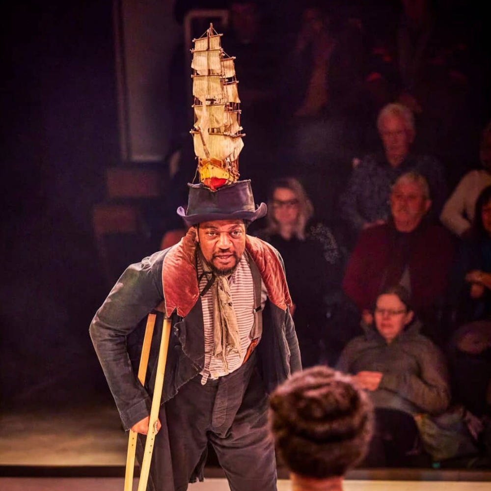 A performer wearing a tall hat with a miniature sailing ship on top, and crutches, performing for an audience in a theater.