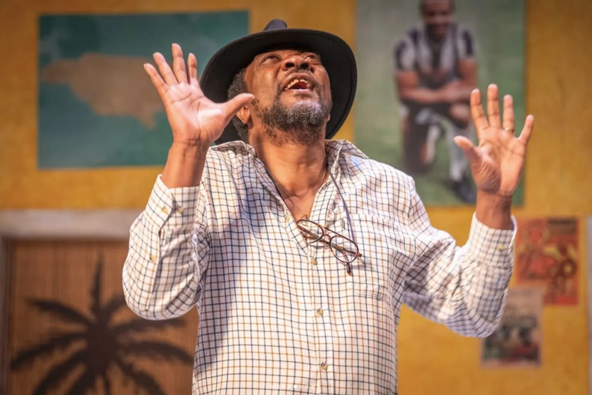 An elderly man with gray hair and beard, wearing a wide-brimmed hat and checkered shirt, gesturing with both hands raised and eyes closed, appearing to be singing or expressing emotion, in a colorful indoor setting with paintings in the background.