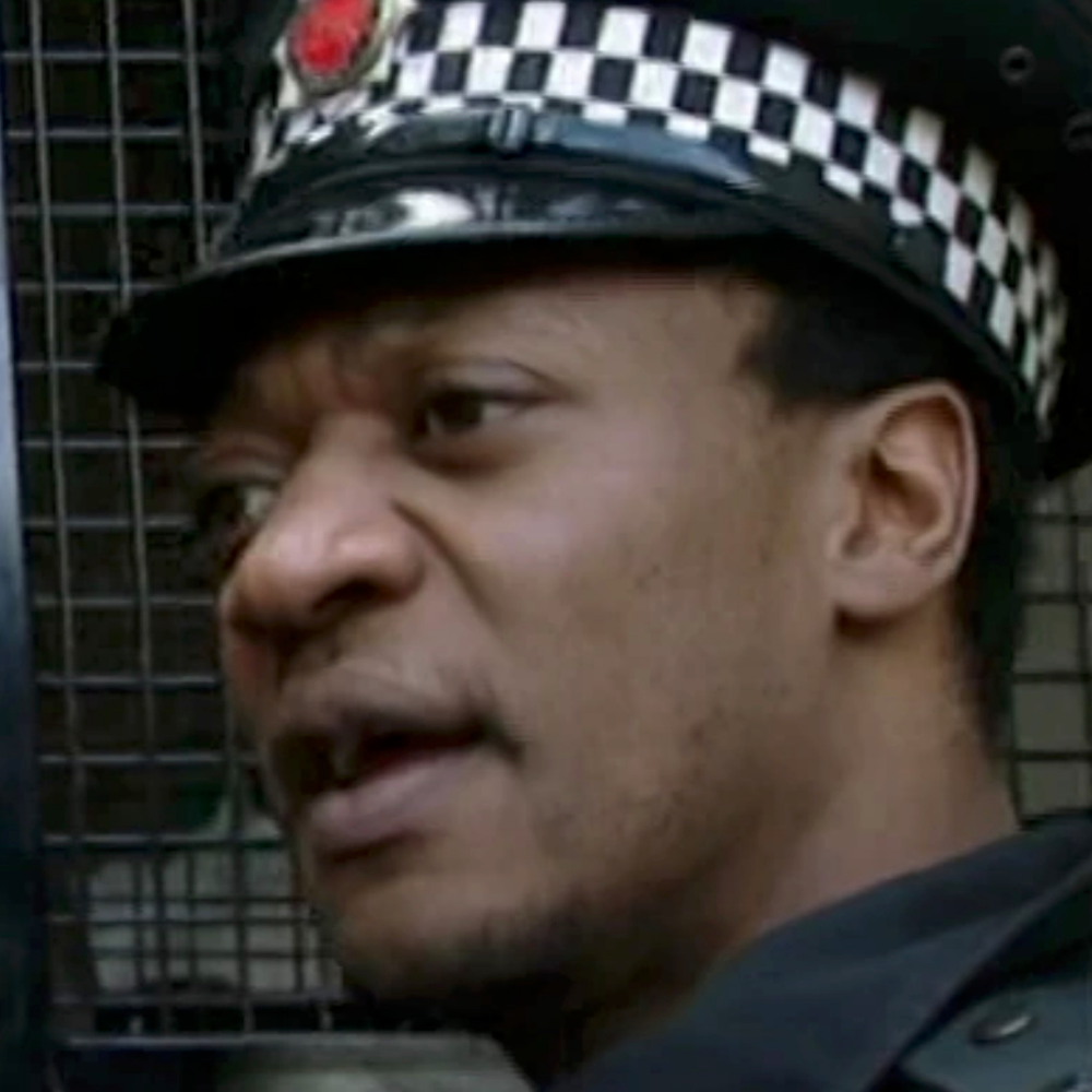 Close-up of a police officer wearing a checkered hat speaking, with a metal grid in the background.