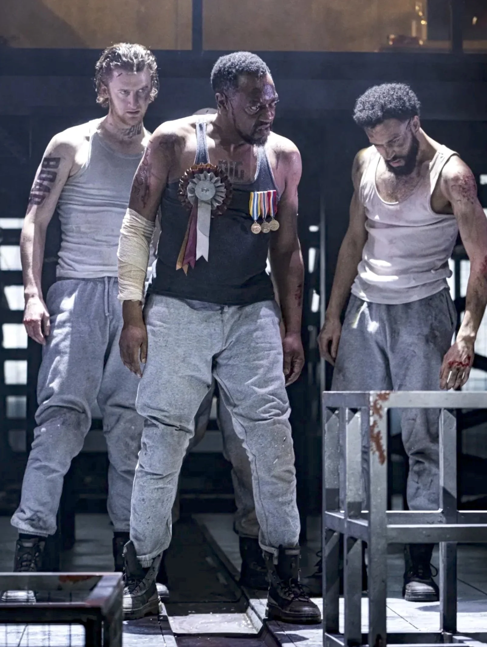 Three men in prison attire stand inside a cell, looking down at a table, with a dark background and metal bars visible.