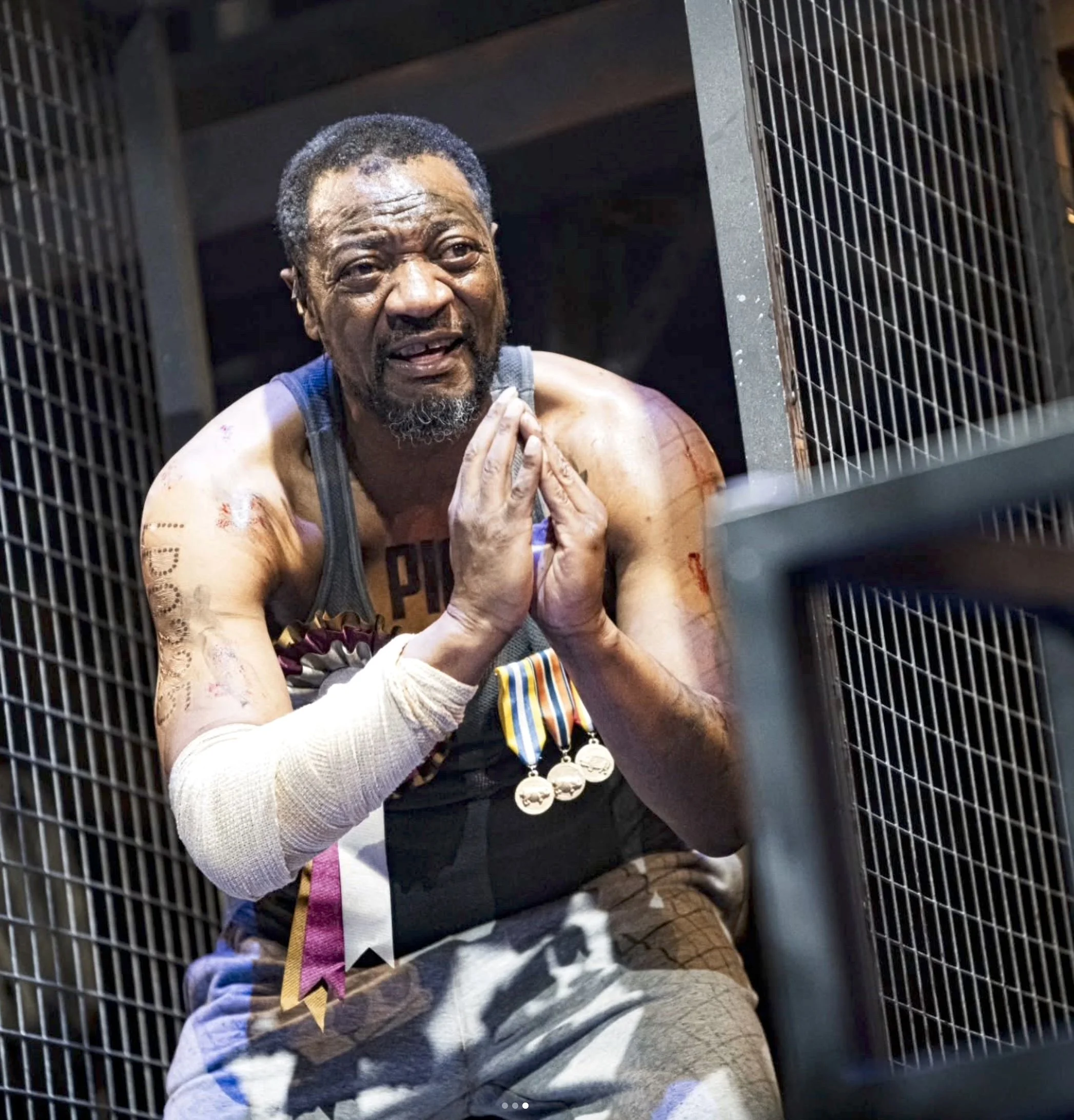 An elderly man with gray hair and a beard sitting inside a black cage or locker. He has a slight smile, is wearing a tank top, and has some bandages on his arms. The man appears to be holding his hands together near his face. The setting looks like a