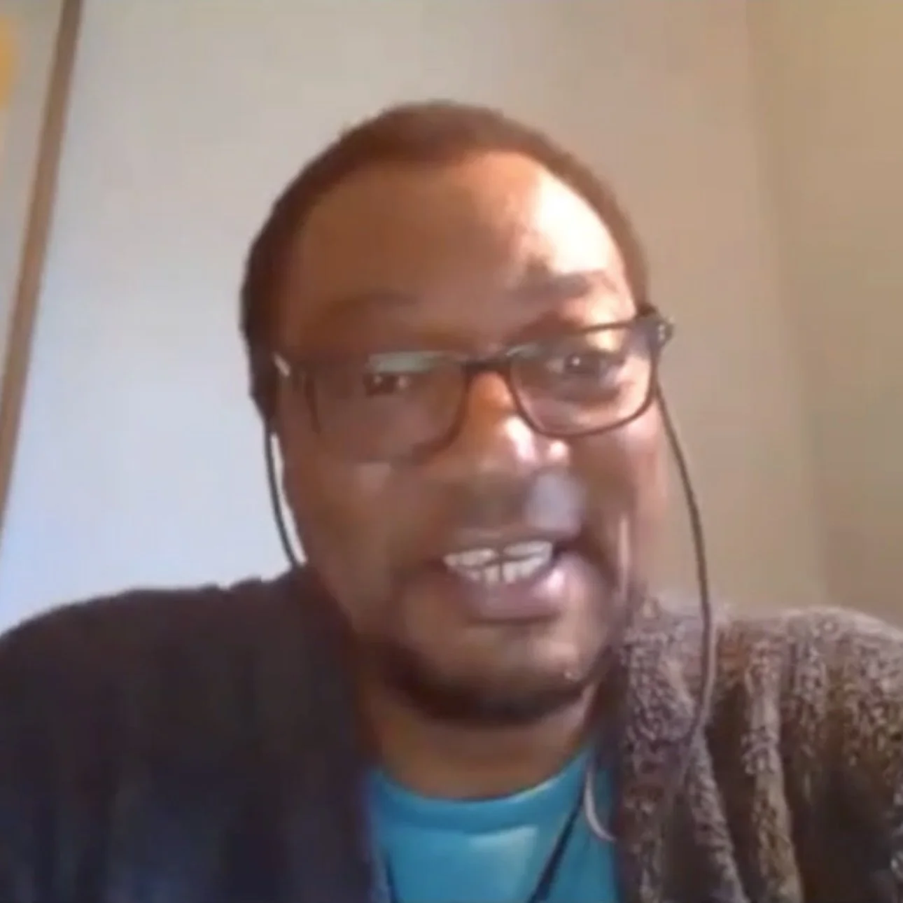 A man wearing glasses and a patterned jacket is smiling indoors. He is wearing a headset with a microphone and has a light blue shirt underneath.