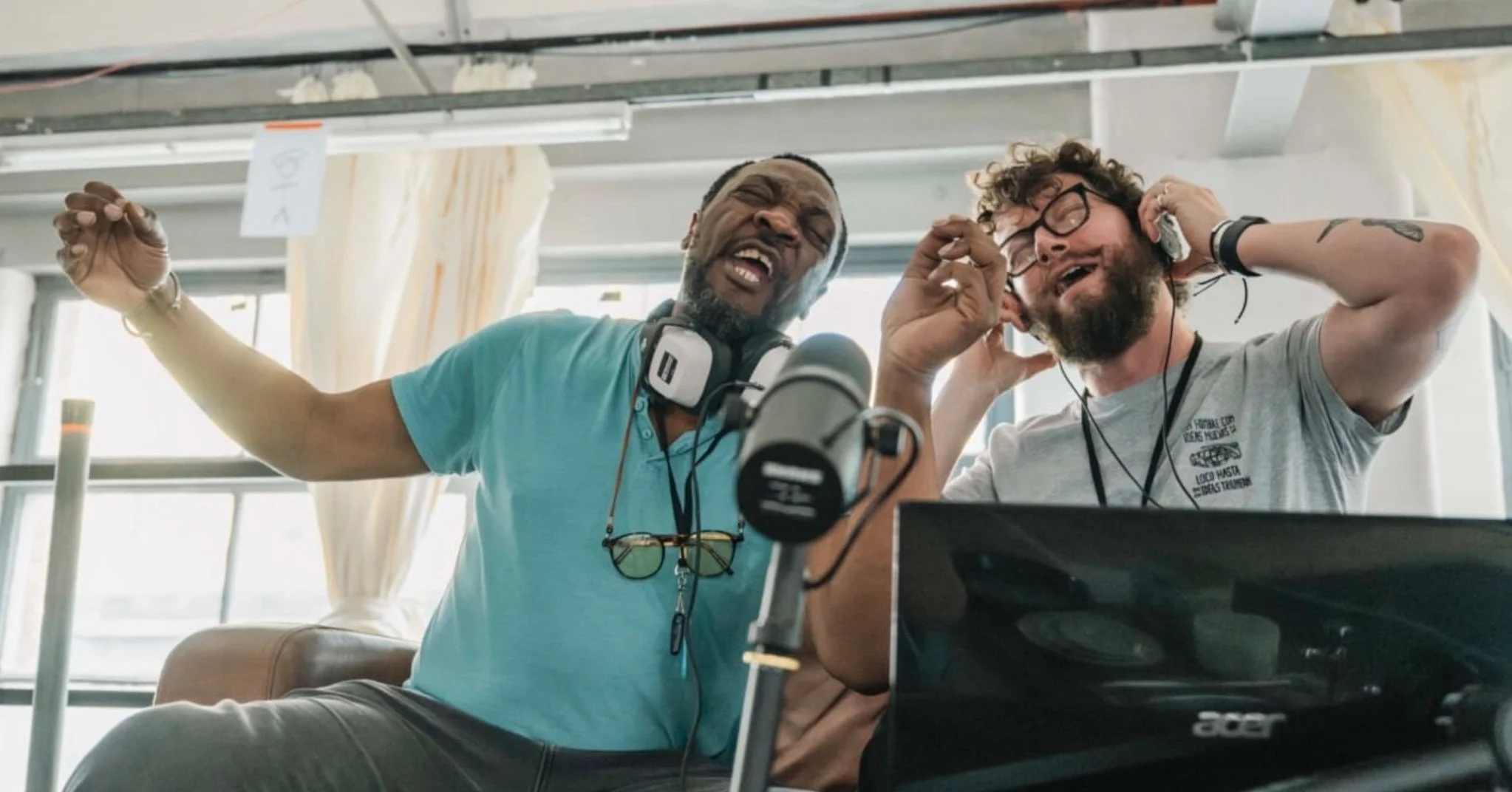 Two men are sitting in a bright room, singing and enjoying music with headphones on. One man is wearing a teal shirt and has a beard, while the other has glasses, a beard, and curly hair. There is a microphone in front of them and a laptop on the tab