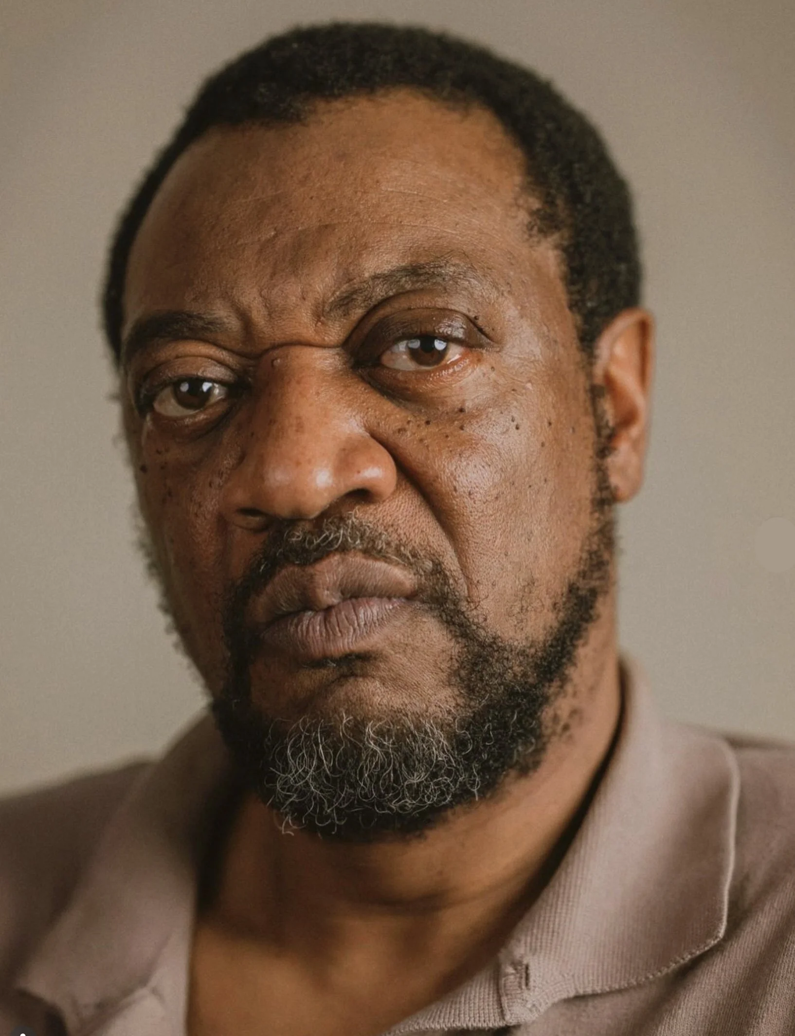 Close-up of a middle-aged man with dark skin, short fractal hair, a beard with gray, and wearing a beige shirt.