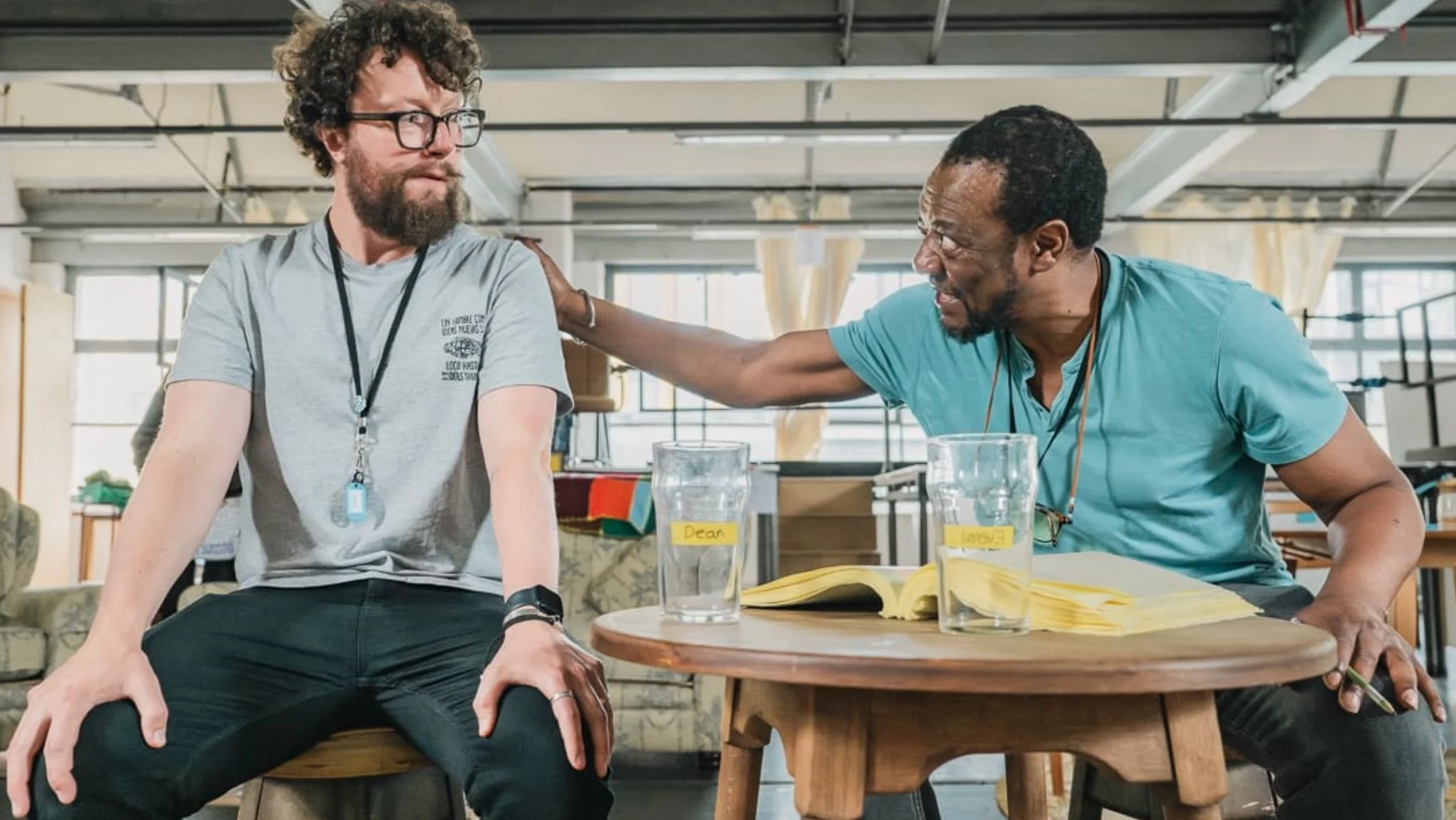 Two men are sitting at a round wooden table in an industrial-style room. One man is African American, wearing a turquoise shirt, glasses, and has a beard. He is smiling and placing his hand on the shoulder of the other man. The other man is Caucasian