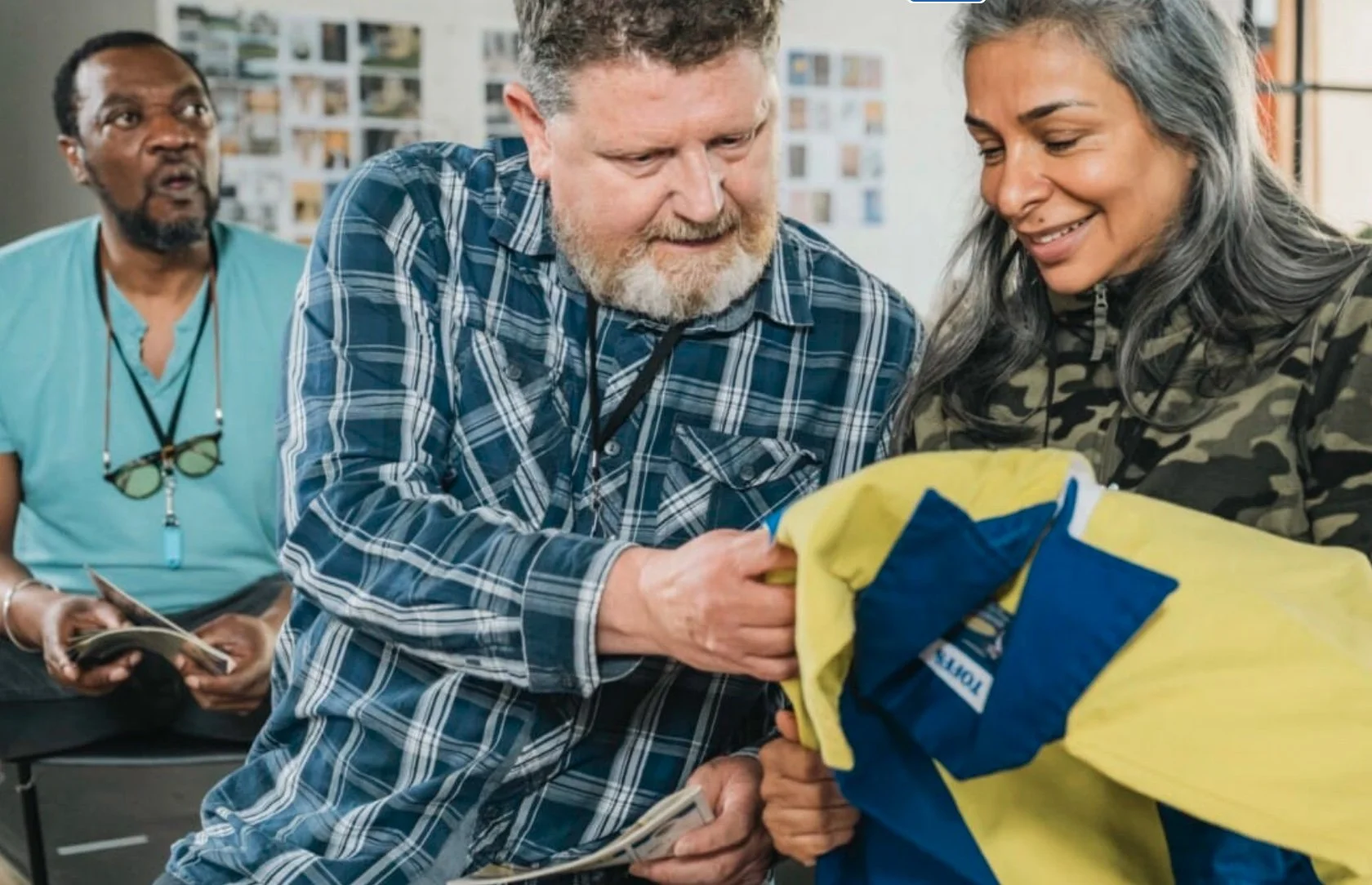 Three people, two men and one woman, are inside a building. The man in the foreground, with a beard and plaid shirt, is handing a yellow and blue fabric to the woman, who has gray hair and is wearing a camouflage jacket. The third person in the backg