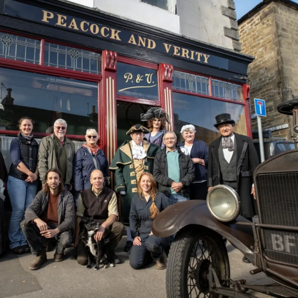 Group of people dressed in period costumes posing in front of a vintage building with a sign that reads "Peacock and Verity." An old car is partially visible in the foreground.