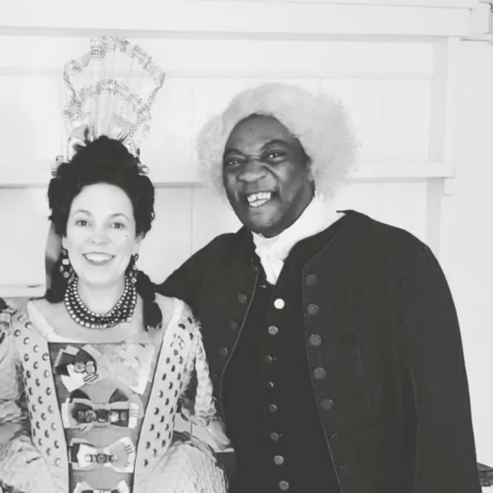 Two people dressed in historical costumes, one person with a large white wig and dark clothing, the other with dark hair, wearing a decorated dress and necklace, standing side by side indoors.