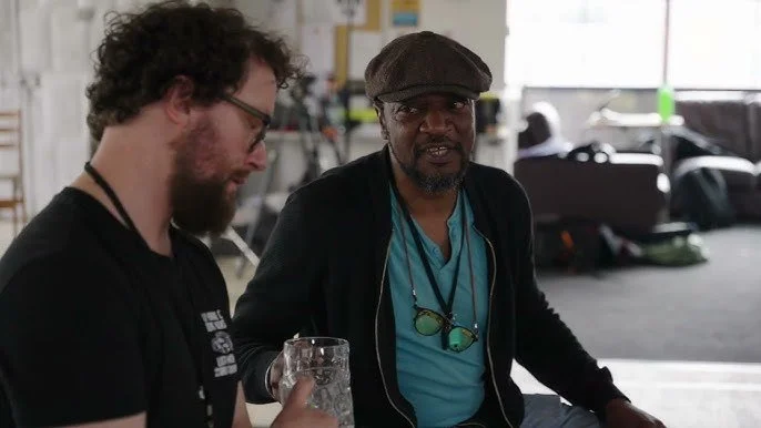 Two men sitting indoors, one with curly hair and glasses holding a glass, the other wearing a cap and sunglasses hanging around his neck, in a casual setting.