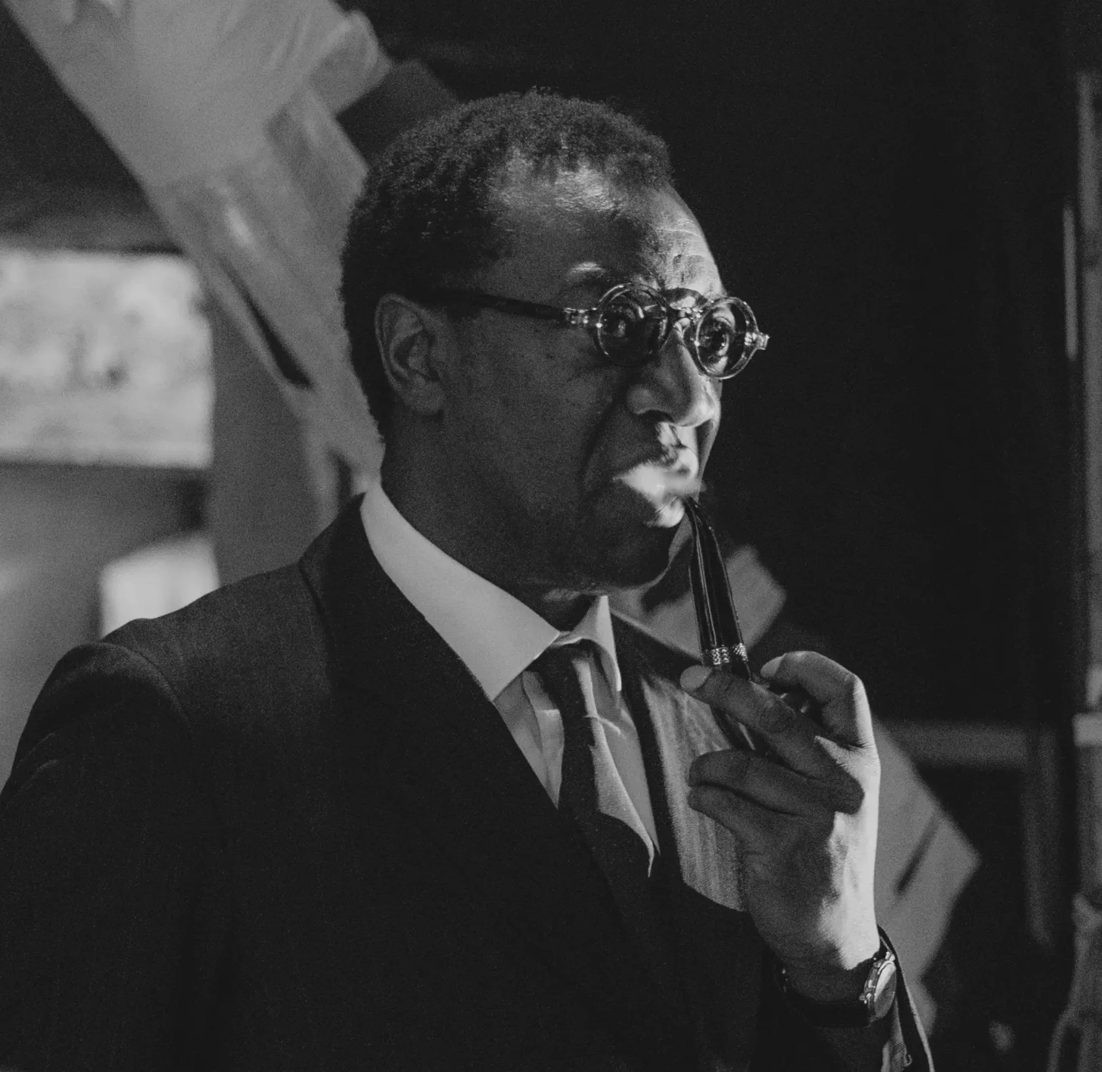 A person with short hair wearing glasses, a suit, and a tie, seen from the side in a black-and-white photo.