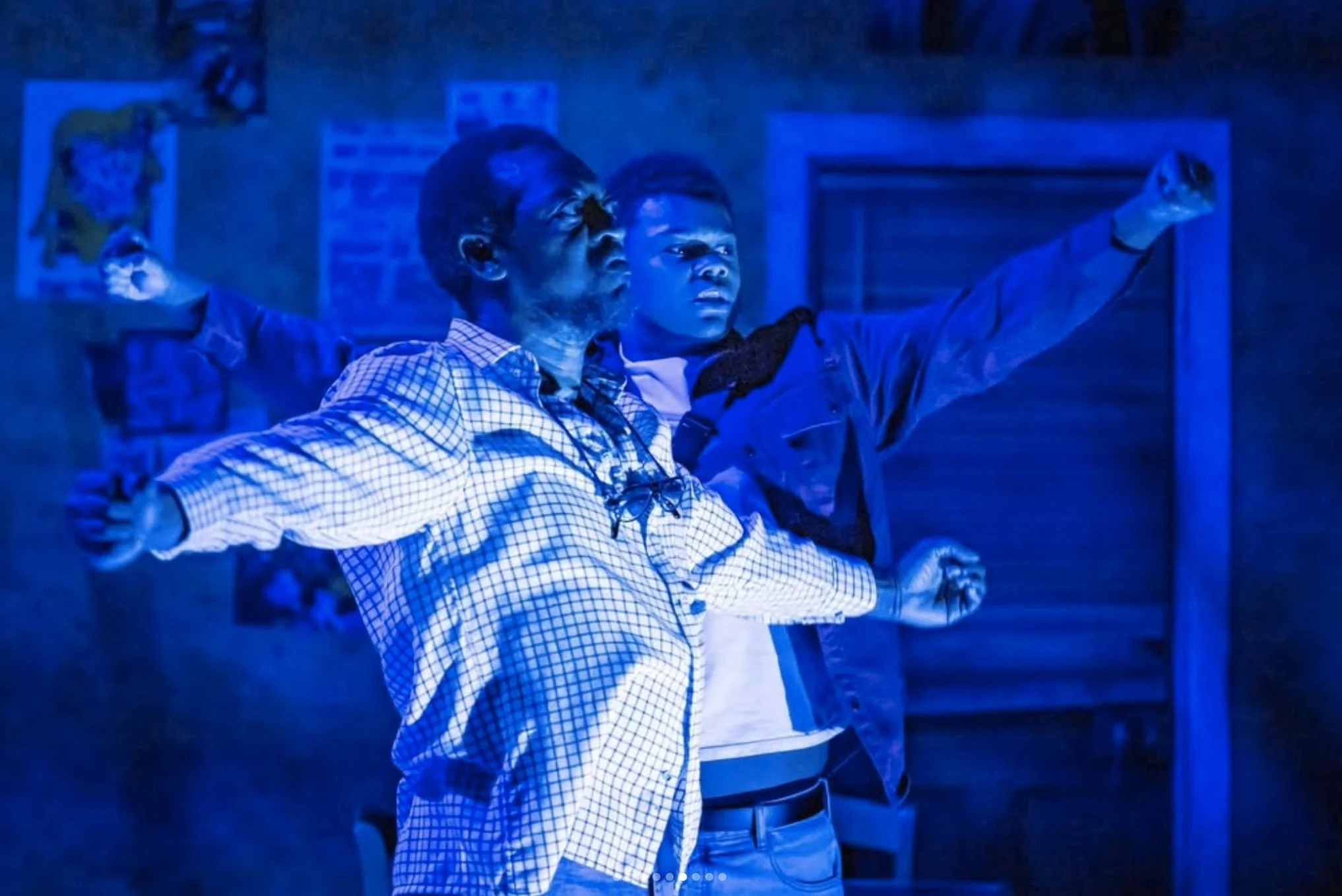 Two young men standing close together in a dark room with blue lighting, one wearing a checkered shirt and glasses, the other in a dark jacket with a choker, both raising their arms in a dance or celebration.