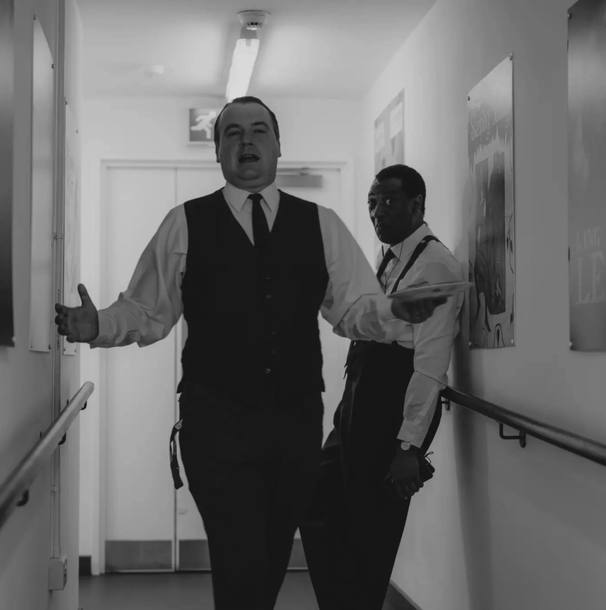 Two men wearing formal attire walking down a corridor, one gesturing with his hand, in black and white photo.
