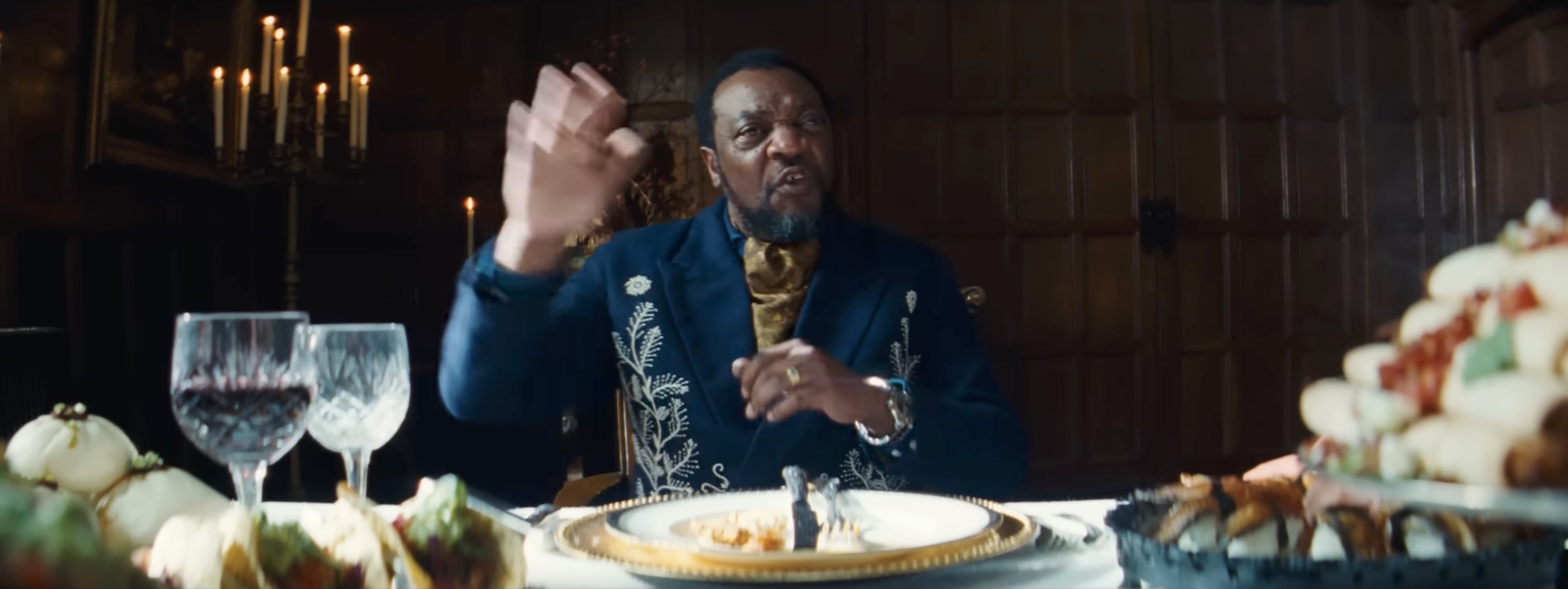 A man in a dark tuxedo with gold embroidery, sitting at a lavish dinner table with wine glasses and dishes, gesturing with his hand inside a dimly lit, wood-paneled room.
